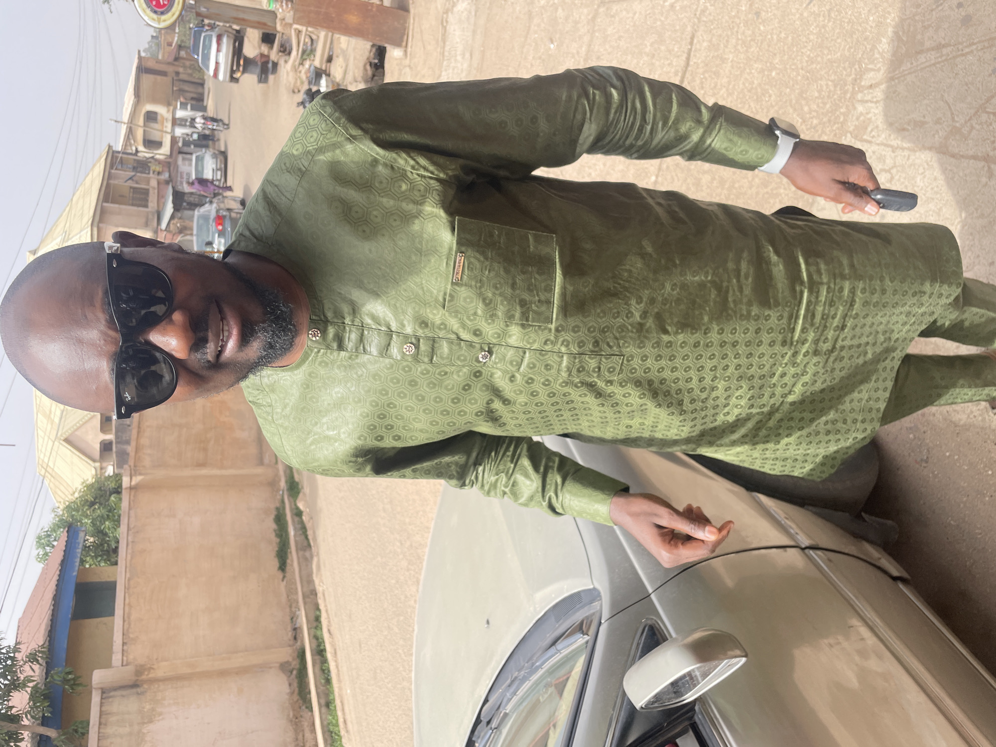 A man in a light green shirt and sunglasses stands outside next to a silver-colored car. The background shows a city street, likely in Jos, Nigeria, given the style of architecture and environmental characteristics. The man is holding an object in his right hand and his left hand is resting on the car. The scene is well-lit, suggesting a sunny day.