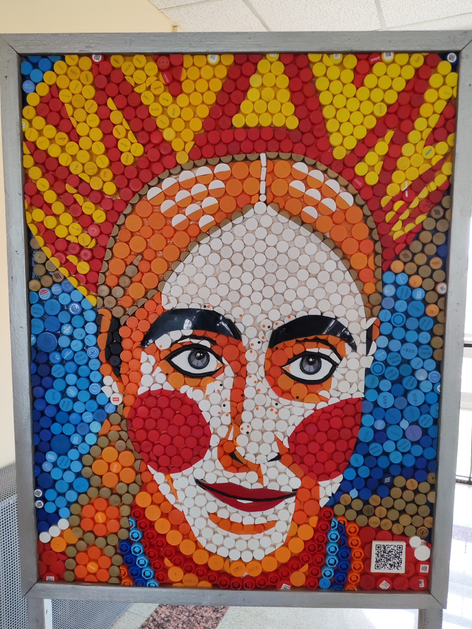 A framed portrait made of plastic bottle caps hangs in what appears to be a public building. The portrait depicts a woman with white skin, red cheeks, dark eyebrows, and large, expressive eyes. Her head is adorned with a headdress made of yellow, red, and orange bottle caps. The background is composed of blue bottle caps. A QR code is located near the bottom right corner of the frame. The frame is a light gray color. The overall setting seems to be indoors near a window. The artwork is located in the city of Agryz, Russia.