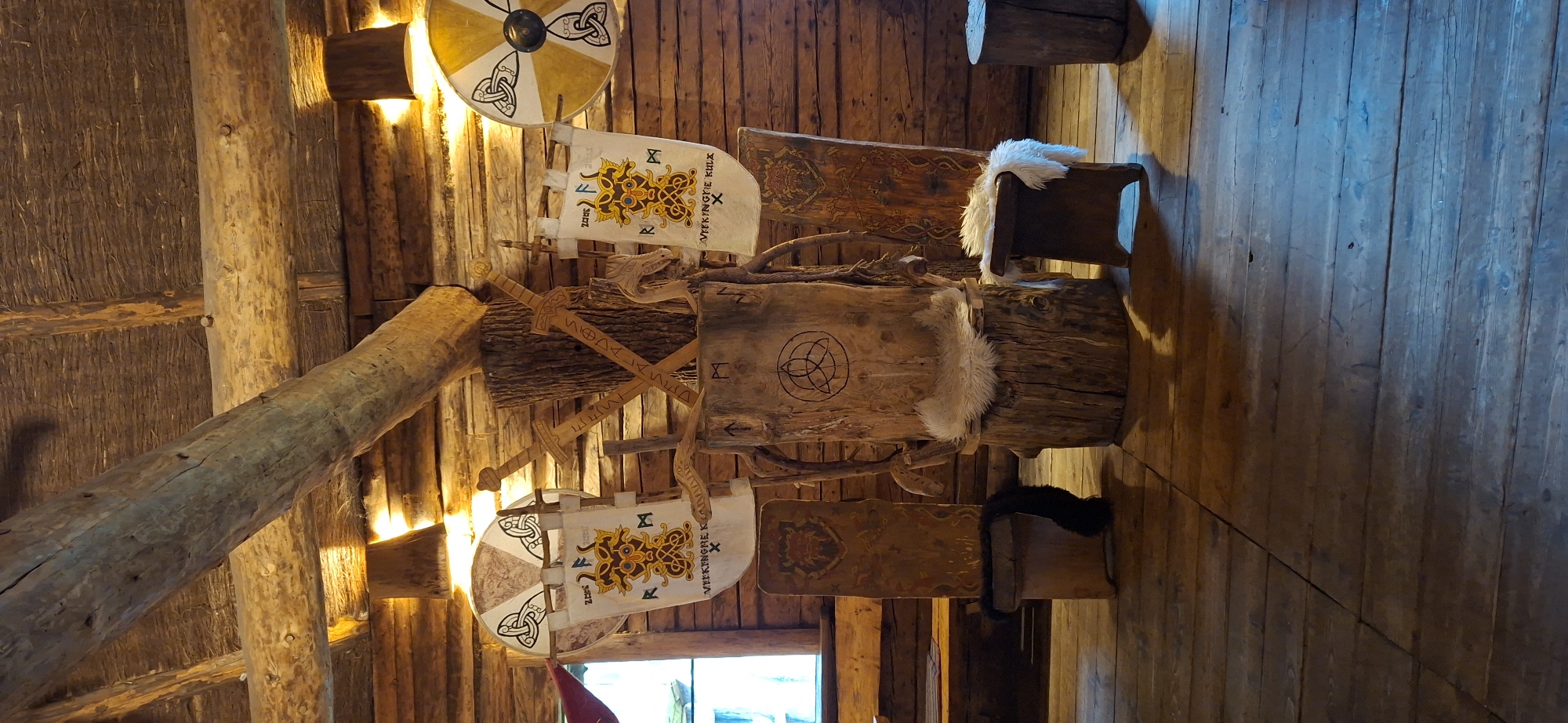 The image depicts a scene set within a rustic, wooden structure, likely a reconstruction or themed exhibit. The environment is characterized by exposed wooden beams on the ceiling and walls constructed from horizontal logs. Lighting appears to be from overhead fixtures, casting a warm glow.
The central focus is a throne-like chair carved from a large tree trunk. It has a rough, natural form, with a circular symbol carved into its backrest. A white, furry pelt is draped over the seat. To the left and right of the throne are two upright wooden panels, likely decorative shields or banners, adorned with intricate patterns and what appear to be runic inscriptions. One panel on the left has a dark fur pelt hanging from its base. In front of the throne, to the right, is a small wooden stool, also topped with a white fur pelt.
Behind the throne, two wooden shields are mounted on the wall, each featuring a circular design with Celtic knotwork. Between the shields and leaning against the throne are two wooden swords crossed over each other, with runic inscriptions carved into their blades. Two banners hang from the wall behind the throne, each depicting a central stylized figure with intricate knotwork and runic lettering.
To the left of the throne, a wooden bench is visible in the background, and a window with wooden shutters can be seen. The floor is made of wide, wooden planks.
The image is likely taken in Saula, Estonia, at a location that is recreating or celebrating Viking or ancient Baltic culture, possibly an open-air museum or a historical reenactment site. No people are present in the image.