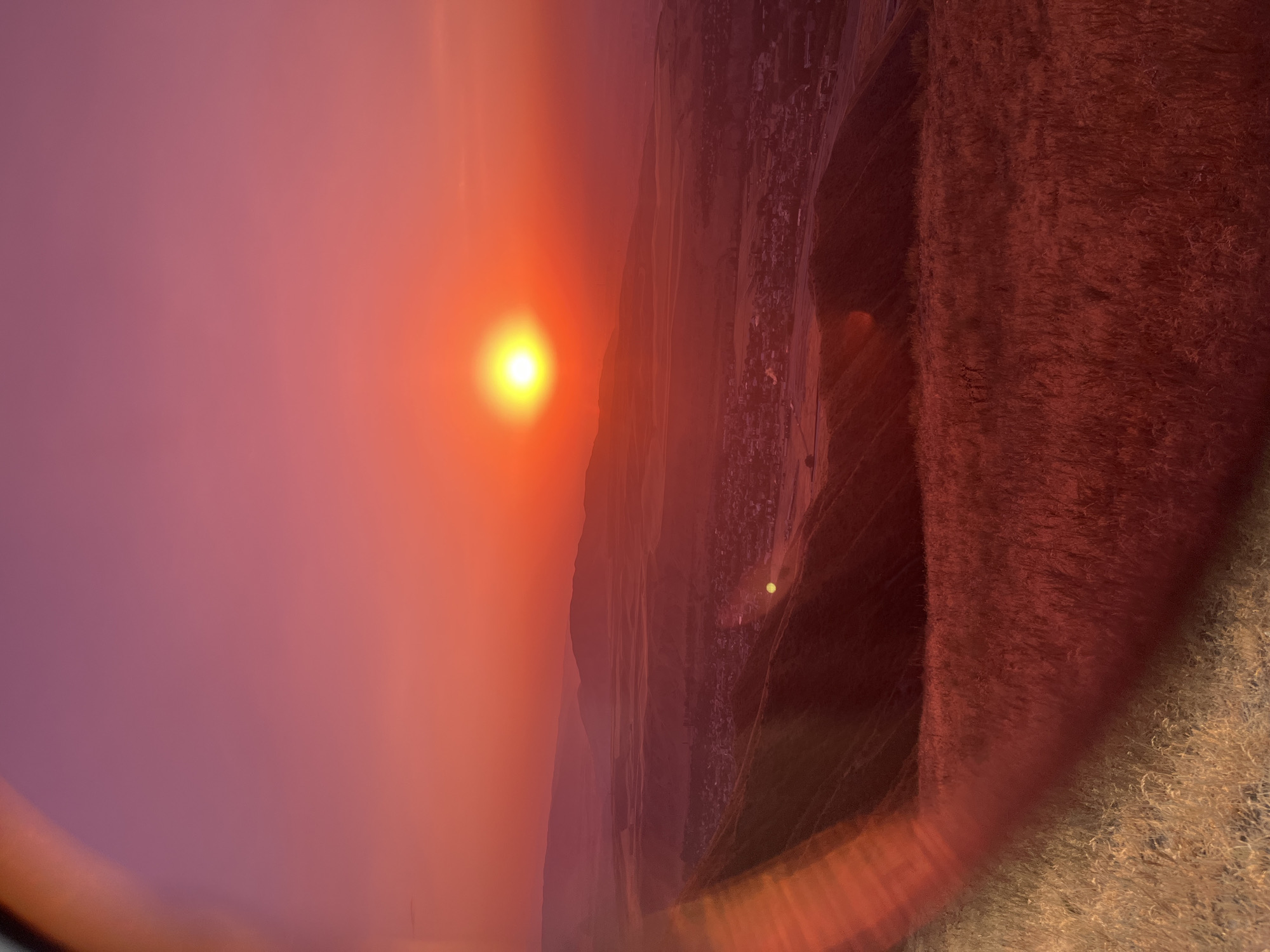 This is a vertical, eye-level, medium shot of a sunset over the city of Besh Kungei, Kyrgyzstan. The sun, a bright yellow orb, is setting in the center of the frame, casting a vibrant orange and pink glow across the sky and landscape. In the foreground, dry, golden grass is illuminated by the setting sun, creating a warm, textured surface. A blurry, reddish-orange lens flare extends from the bottom left corner of the image. In the midground, rolling hills are visible, leading down to a town nestled in a valley. The houses and buildings of the town are discernible, with a hint of lights appearing to be on. In the background, distant mountains are silhouetted against the colorful sky, their shapes softened by the atmospheric haze. The overall impression is one of natural beauty and tranquility.
