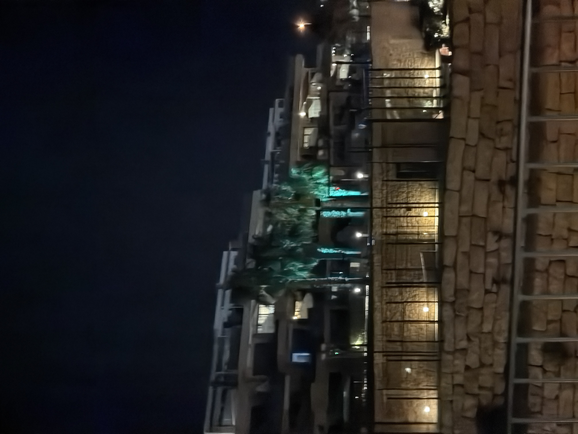 The image shows a multi-story modern building with balconies and illuminated windows at night. Several palm trees are visible in front of the building, some of which are lit with green lights. A stone wall and metal fencing are in the foreground, with more lights illuminating the wall. The sky is dark, indicating nighttime. The scene is consistent with a resort or residential complex in a warm climate, possibly located in Finestrat, Spain, given the presence of palm trees and modern architecture often found in such Spanish coastal towns.