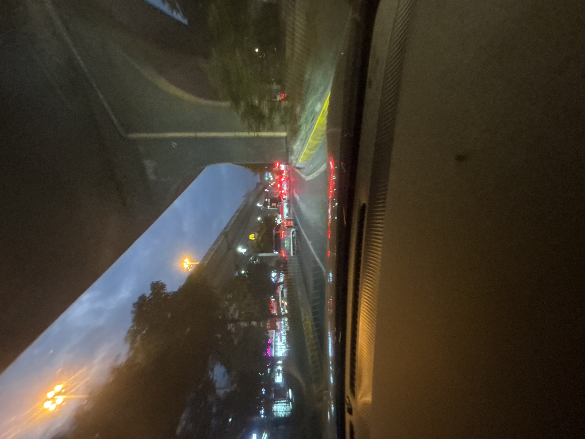 A first-person perspective shot taken from inside a vehicle in Baiyun District, China, captures a nighttime urban scene under an overpass. The sky is a dusky blue, indicating twilight or early night. Streetlights and lights from businesses illuminate the surroundings. On the left, a row of buildings with vibrant neon signs is visible. To the right, the concrete structure of an overpass dominates the upper portion of the frame. Several cars are on the road, with their taillights creating streaks of red. The road surface appears wet, suggesting recent rain. A golden arch logo, likely for McDonald's, is visible on a building in the middle distance. The foreground of the image is dominated by the dashboard of the vehicle, with a textured surface.