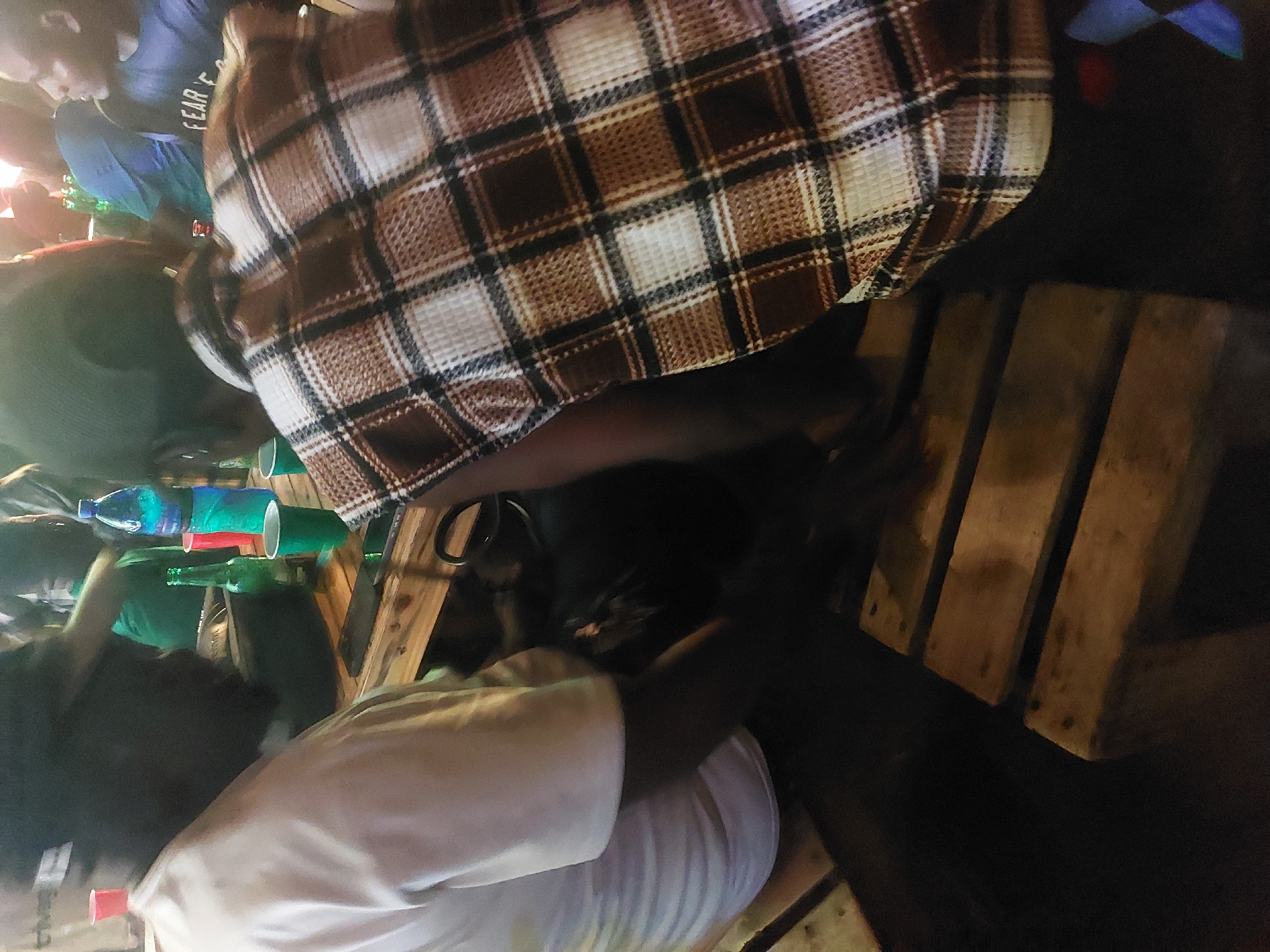 The image shows a dimly lit gathering of people around a wooden pallet table. Several individuals are visible, dressed in casual attire, including a plaid shirt and a white shirt. There are bottles and green cups on the table, possibly containing drinks. The setting appears to be an informal outdoor space, possibly in a neighborhood or public area. The location is described as being in Dar es-Salaam, Tanzania.