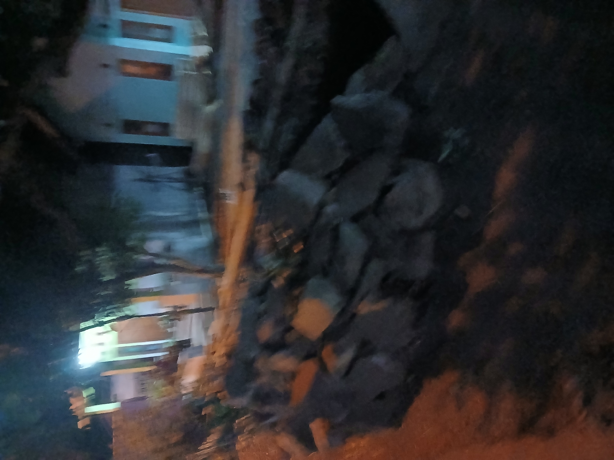 The image depicts an outdoor scene at night, likely in the city of Kademangan Kulon, Indonesia. In the foreground, a pile of rough-hewn stones or concrete debris is visible. Beyond this debris, a light-colored house with dark-trimmed windows and doors is situated. The house appears to be two stories with a pale blue exterior. Trees with dark foliage are present in front of and around the house, casting shadows. The overall lighting suggests artificial illumination from within the house or from streetlights, creating a dim and somewhat blurry atmosphere. The ground in front of the house appears to be a dirt or gravel surface. The image lacks discernible human activity.