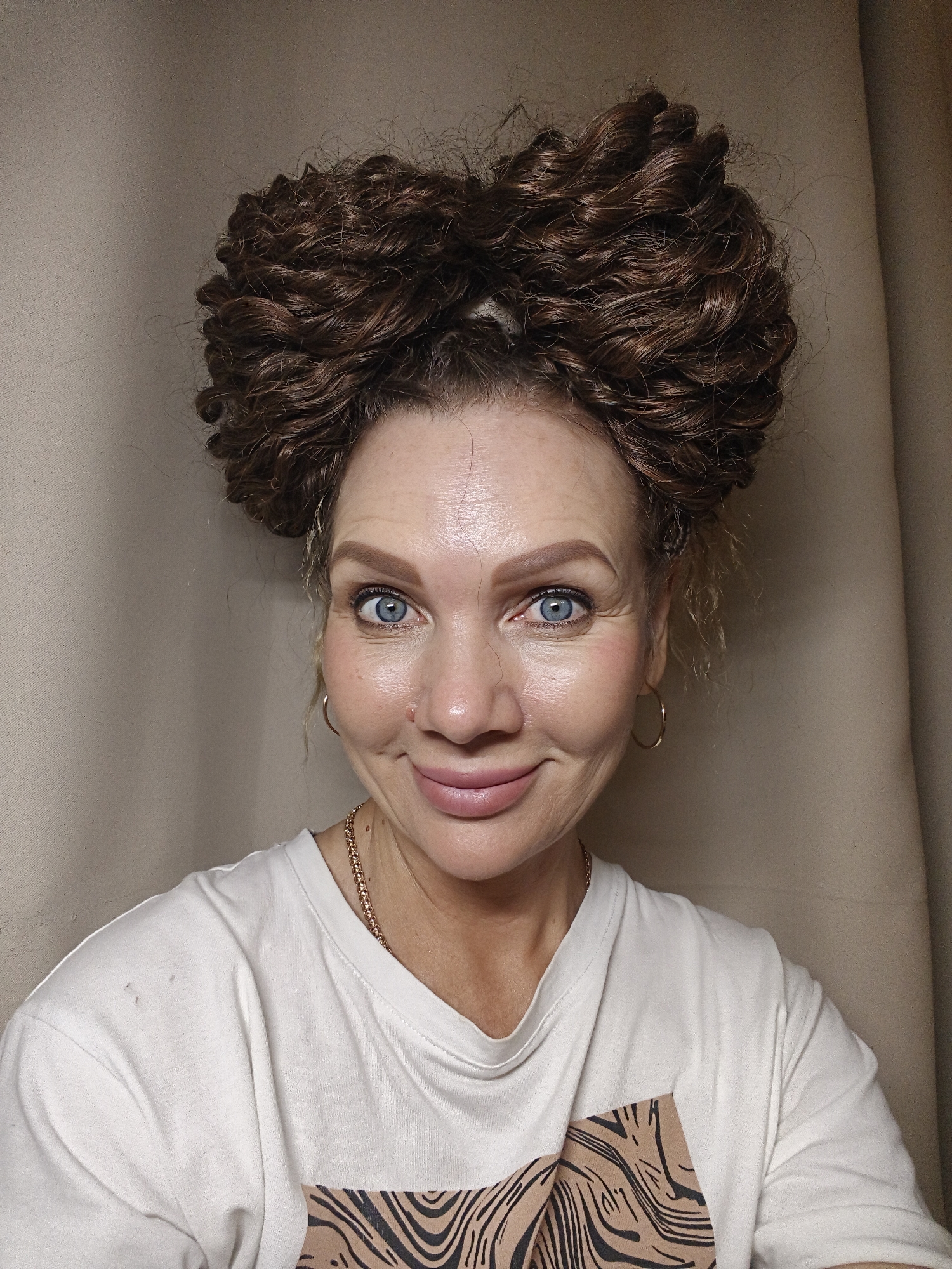 An indoor portrait displays a middle-aged woman with light skin and blue eyes, smiling directly at the viewer. Her dark brown, curly hair is styled into two voluminous coiled buns on top of her head, forming a heart-like shape. She wears subtle makeup, including blush and light pink lip gloss, and has a small gold stud in her left nostril. Her accessories include gold hoop earrings and a gold chain necklace. She is dressed in an off-white t-shirt featuring a brown and black abstract or animal print graphic on the lower right chest area, and two small dark marks are visible on her left shoulder. The background consists of a plain, light beige or tan wall or curtain, indicating an indoor setting in the city of Unknown, Unknown.