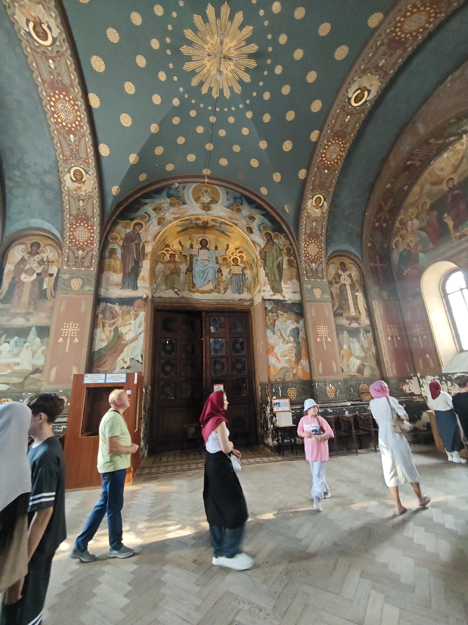 The image captures the interior of a large, ornately decorated building, likely a religious structure such as a church or cathedral, viewed from a wide-angle perspective looking upwards. The ceiling is a dark teal-green, adorned with numerous golden, circular, floral motifs and a central golden sunburst design. Arched structural elements are prominent, featuring intricate patterns and framed religious figures with halos. The walls are extensively covered with detailed murals depicting various religious scenes and figures, executed in a style reminiscent of Byzantine or Eastern Orthodox iconography, with rich color palettes.
In the central background, a large, dark wooden double door with glass panels is visible, flanked by more murals. A tall, arched window on the right side illuminates the space, casting strong light and distinct shadows on the floor. The flooring is light-colored wooden parquet, laid in a herringbone pattern.
Approximately eight to ten individuals are present in the lower half of the frame, observing the interior. From left to right: a person in a light gray hijab and another in a dark short-sleeved top with white arm stripes are partially visible. A man with light hair, wearing a light green polo shirt, blue jeans, and gray sneakers, walks towards the right. A woman in a red hijab, white top, and dark skirt walks towards the right. Another woman in a white polka-dotted dress and a pink hijab walks away from the camera, carrying a light-colored handbag. A young girl in a pink t-shirt, light blue pants, and a light-colored bucket hat stands facing inward. Additional figures are partially visible in the mid-ground on the right. Individuals are either walking or standing, observing the decorated environment. The setting is indoors, in the city of Unknown, Unknown.