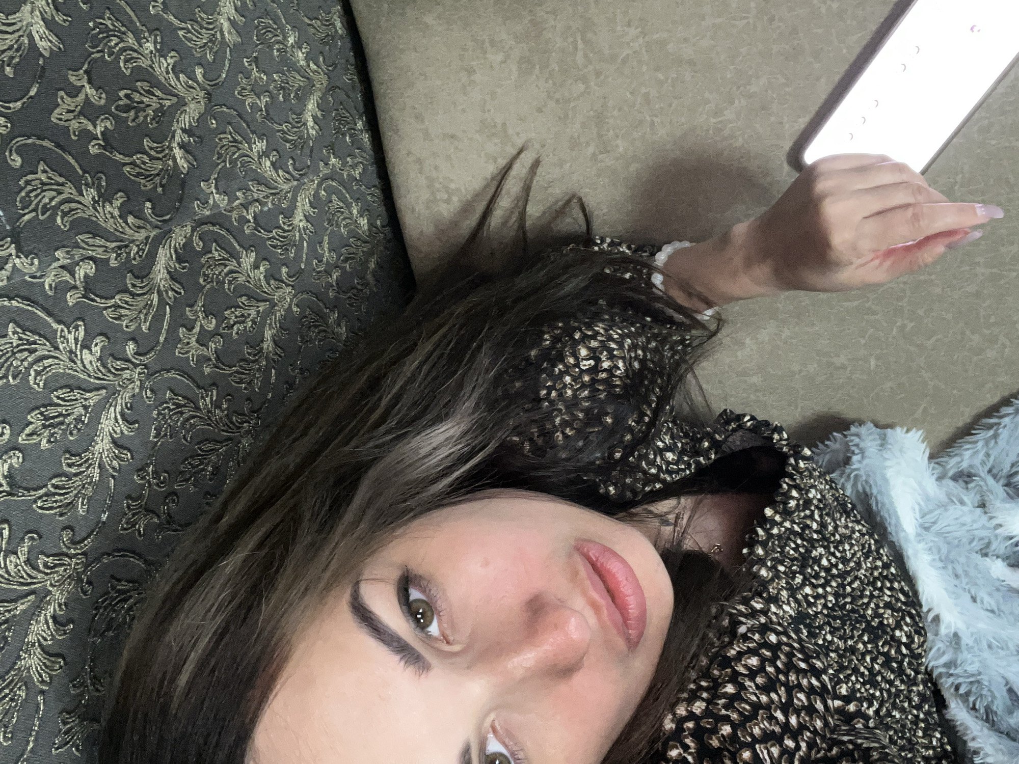 A young woman with brown hair and green eyes is lying down on a beige sofa, possibly in a living room. She is wearing a black and gold patterned top. Her left arm is resting on the sofa, and her right hand is holding a white smartphone. The background is a dark gray patterned fabric, possibly a wall or headboard. A light blue fuzzy blanket is visible on the sofa next to her. The lighting suggests it may be indoors, with a light source coming from the smartphone.