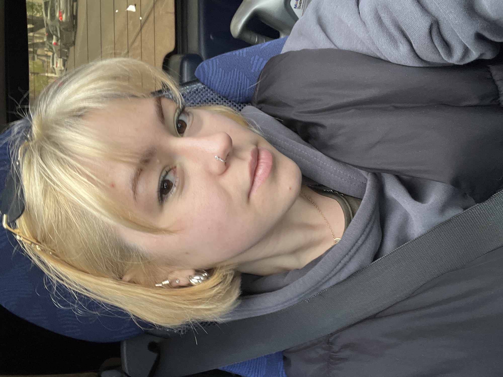The image displays a rotated medium close-up of a young person with blonde hair, looking directly into the camera. They are seated in what appears to be a vehicle, possibly a bus or train, indicated by the blue textured fabric of the seat and a dark grey seatbelt running across their torso.
The person has fair skin, dark eyes, and a neutral, somewhat pensive expression. They sport a small, ornate hoop piercing in their right nostril and multiple small hoop earrings or ear cuffs on their left ear. Their blonde hair is styled with bangs that frame their face.
They are wearing a grey hooded sweatshirt layered under a dark grey or black puffer vest. A delicate gold chain necklace with a small circular pendant is visible around their neck. In the background, behind their right shoulder, a grey plastic surface has a white sticker with visible text that reads "Mobil Super". Through a window, out-of-focus reflections of what seem to be urban elements, including some distant red lights, are discernible. The lighting is soft and diffused.