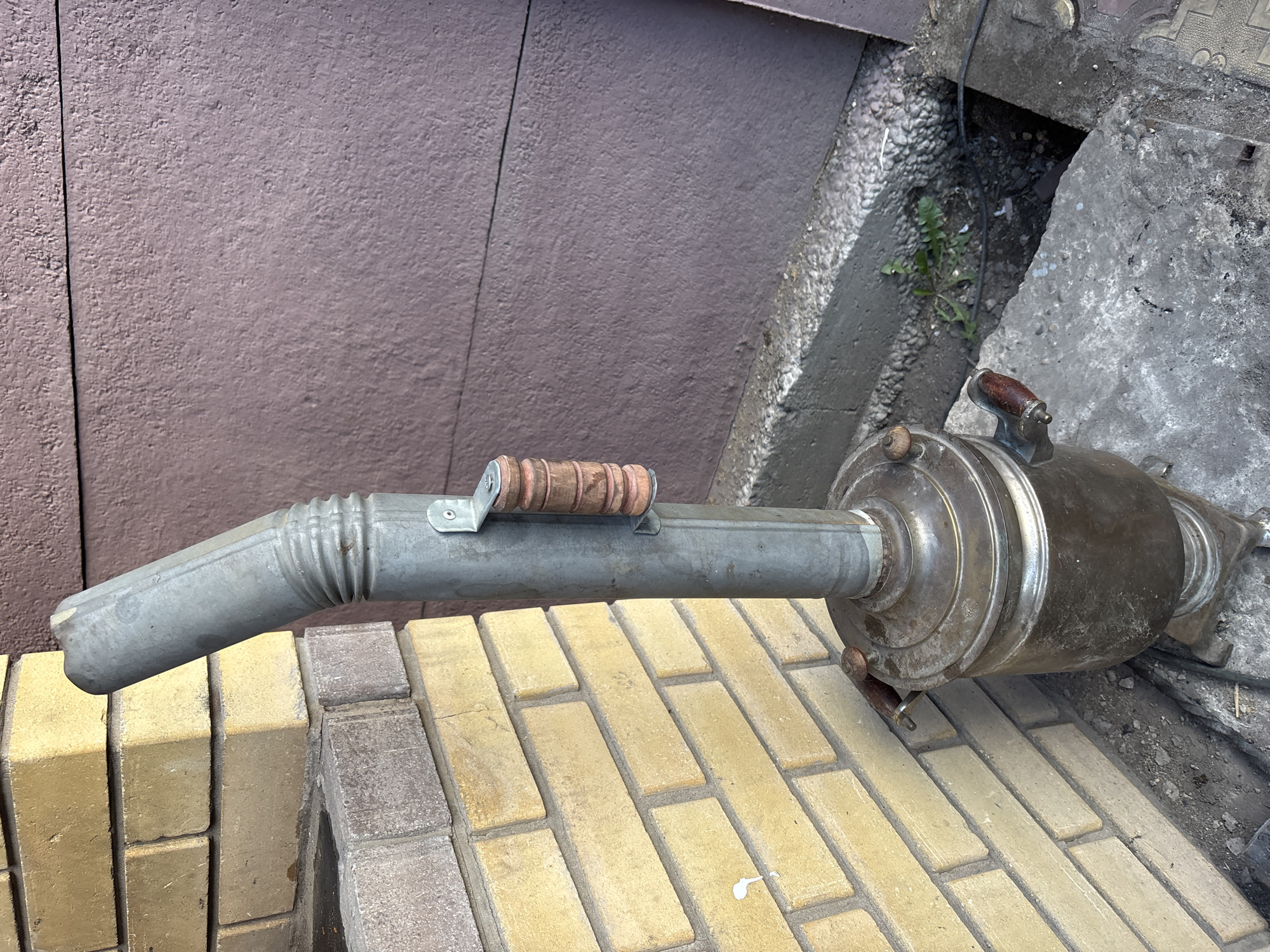 A weathered, metal samovar with wooden handles stands on a concrete surface next to a yellow brick wall. A grey metal pipe, likely a chimney or vent, extends upwards from the top of the samovar and bends to the left. The pipe is secured to the samovar by a metal bracket with a wooden handle. The background consists of a textured, dark brown wall. The ground around the samovar is a mix of dirt, small stones, and some concrete debris. There are no people visible in the image. The setting appears to be an outdoor urban environment, possibly an alleyway or backyard in Chernogorsk, Russia.