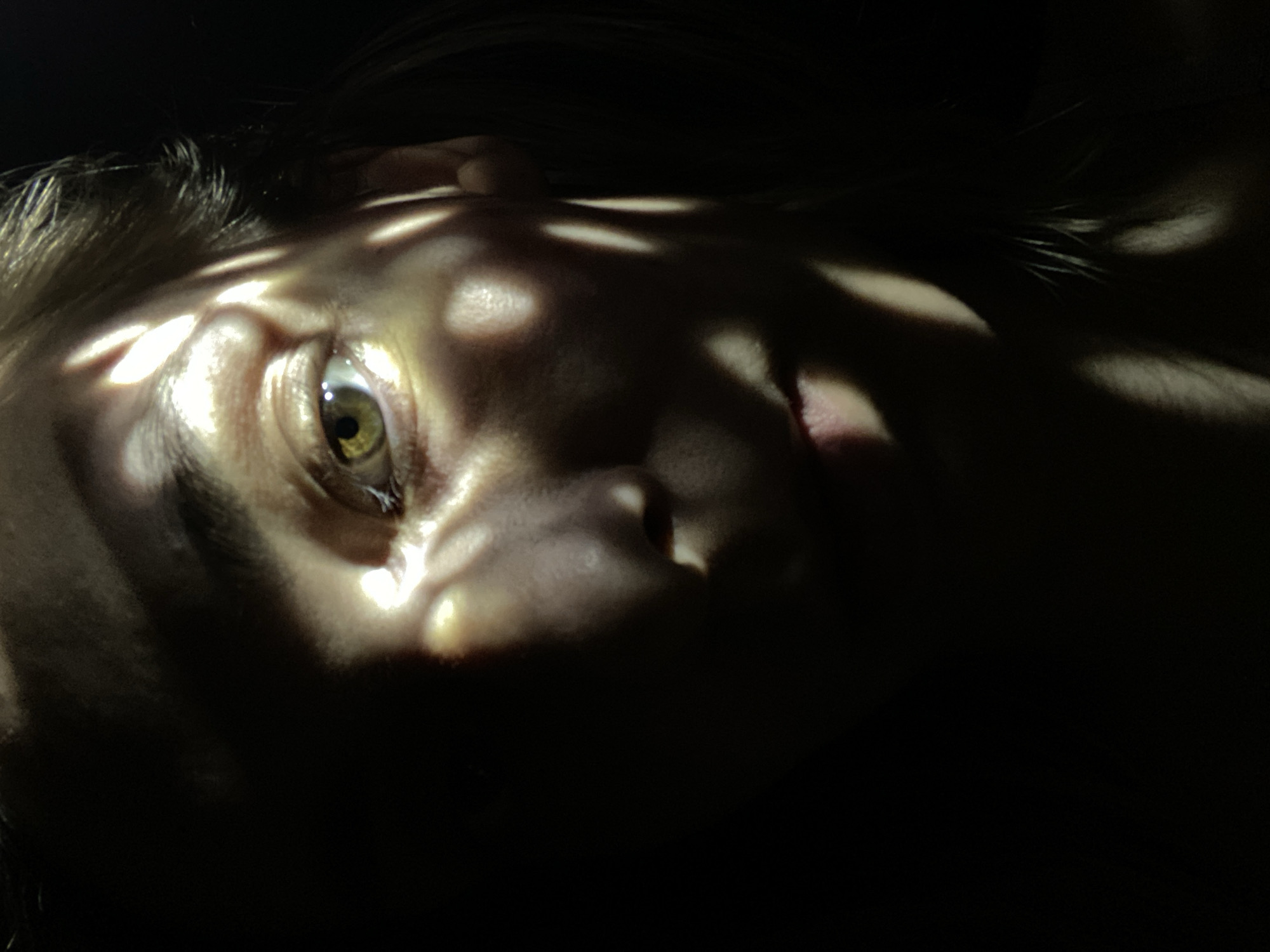 A close-up, slightly angled shot of a person's face in near darkness, illuminated by dappled light. The person is likely lying down, with their head slightly tilted towards the viewer. The light creates a pattern of shadows and highlights across their face, emphasizing the textures of the skin. The person's eye is open and directed towards the light. The background is entirely obscured by darkness. There is no information available within the image itself to indicate the location as Chaikovsky, Russia.