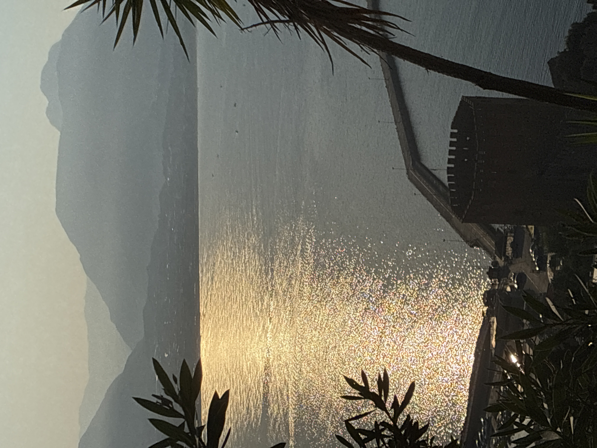 The image depicts a scenic view overlooking the Mediterranean Sea at sunset, likely from a vantage point on the hill of Alanya Castle in Alanya, Turkey. The sun's reflection creates a shimmering, golden path across the water. In the foreground, dark silhouettes of olive branches and palm fronds frame the scene. Below, a portion of the historic Red Tower and a long pier extending into the sea are visible. In the distance, hazy mountains form a layered backdrop against the soft twilight sky. Several small boats are scattered across the water. The overall atmosphere is tranquil and serene.