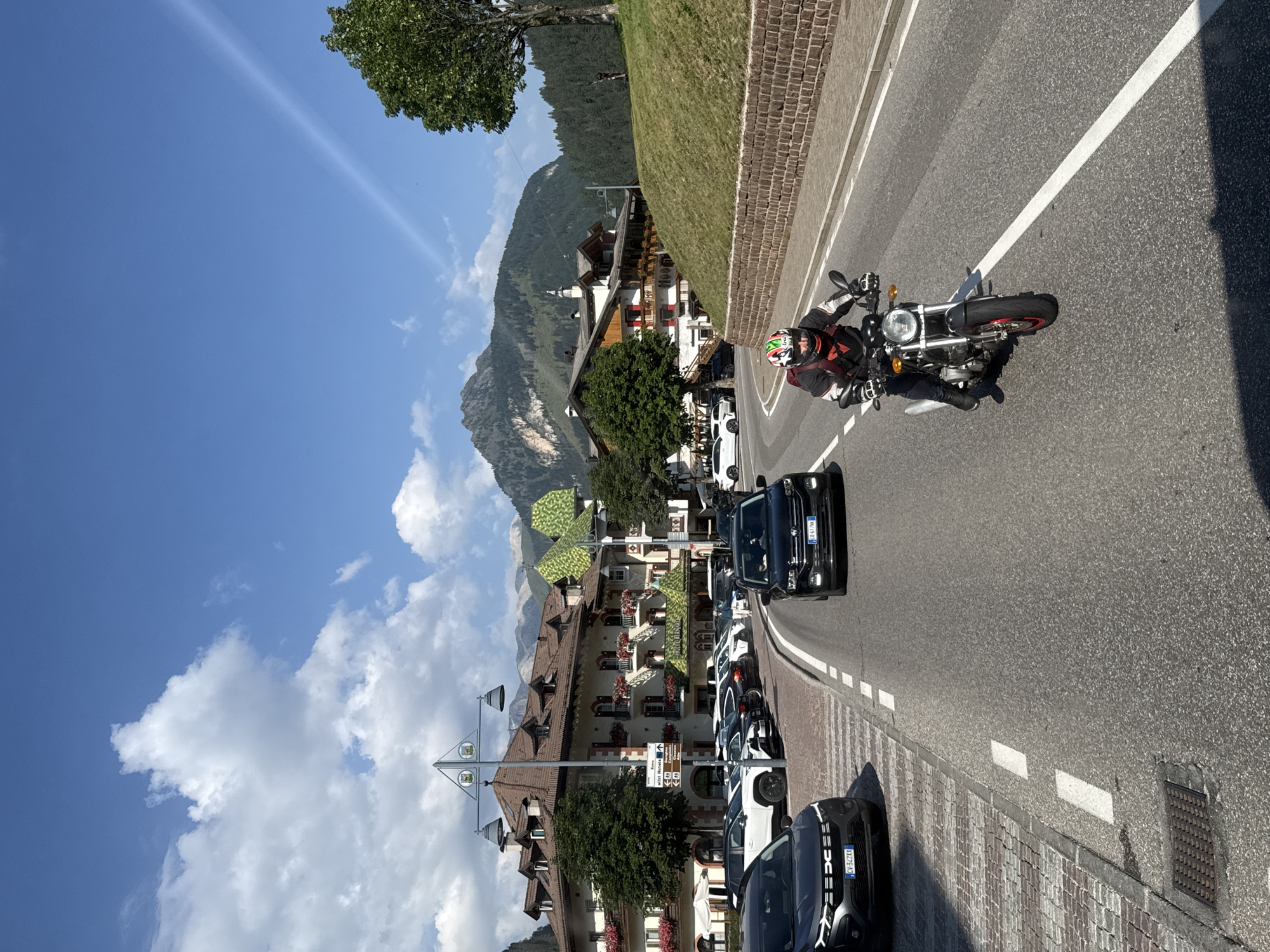 The image depicts a street scene in Canazei, Italy, on a sunny day with scattered clouds. In the foreground, a person wearing a motorcycle helmet and riding gear is on a motorcycle, traveling on a paved road. The motorcycle has a red rim on its front wheel. To the left of the road, several cars are parked along a sidewalk. Beyond the parked cars, a large, multi-story building with a distinctive brown roof and numerous windows is visible. This building appears to be a hotel, with a sign that reads "HOTEL GARDENA." Further in the background, rolling hills covered in trees rise towards a mountainous peak. The sky is bright blue, with some fluffy white clouds and a faint contrail. The overall environment suggests a mountain resort town.