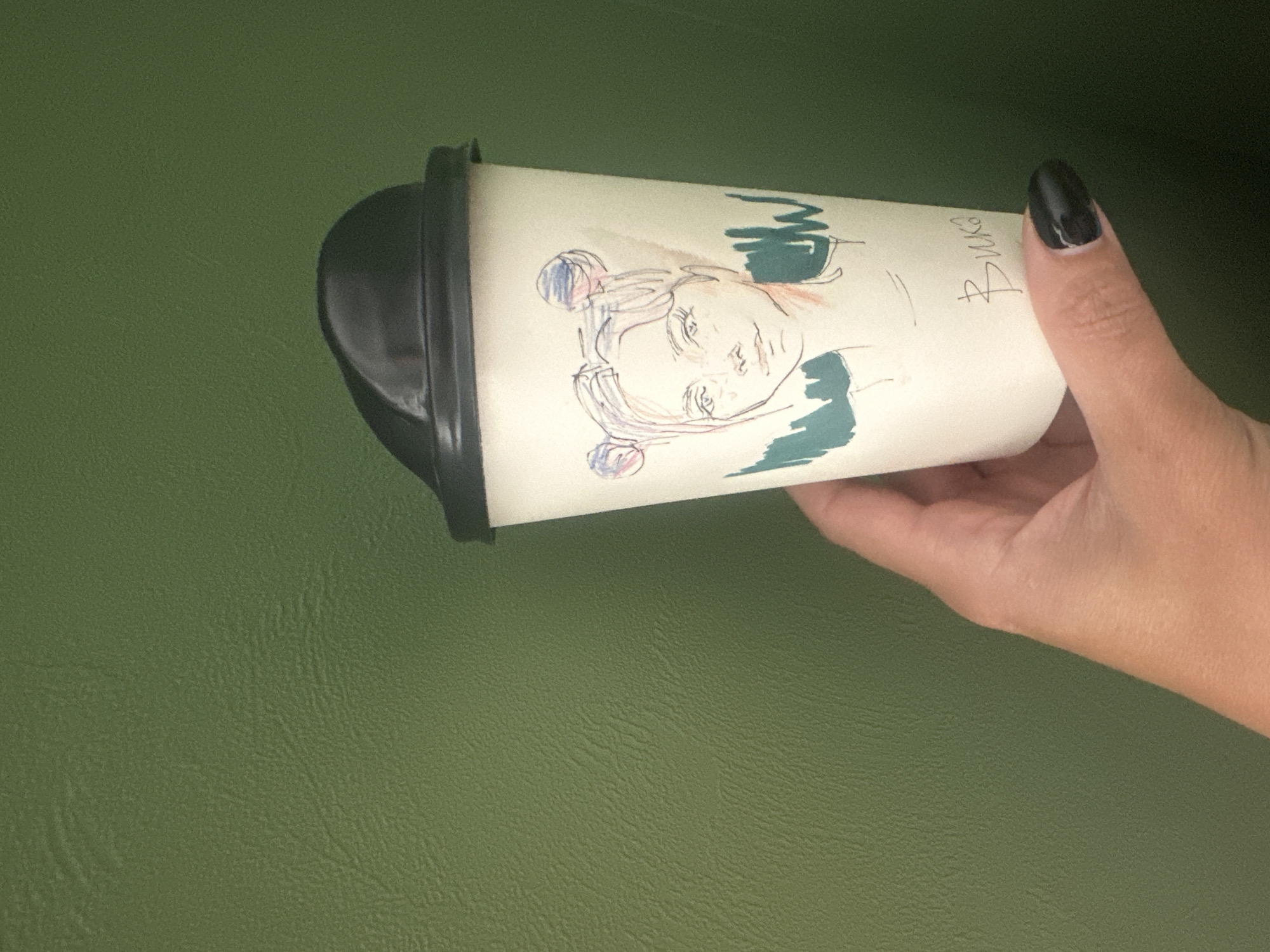 A hand with a black fingernail holds a white paper coffee cup with a black lid. The cup has a sketch of a face and the name "Вика" written on it. The background is a textured green surface. The location cannot be determined from the image.