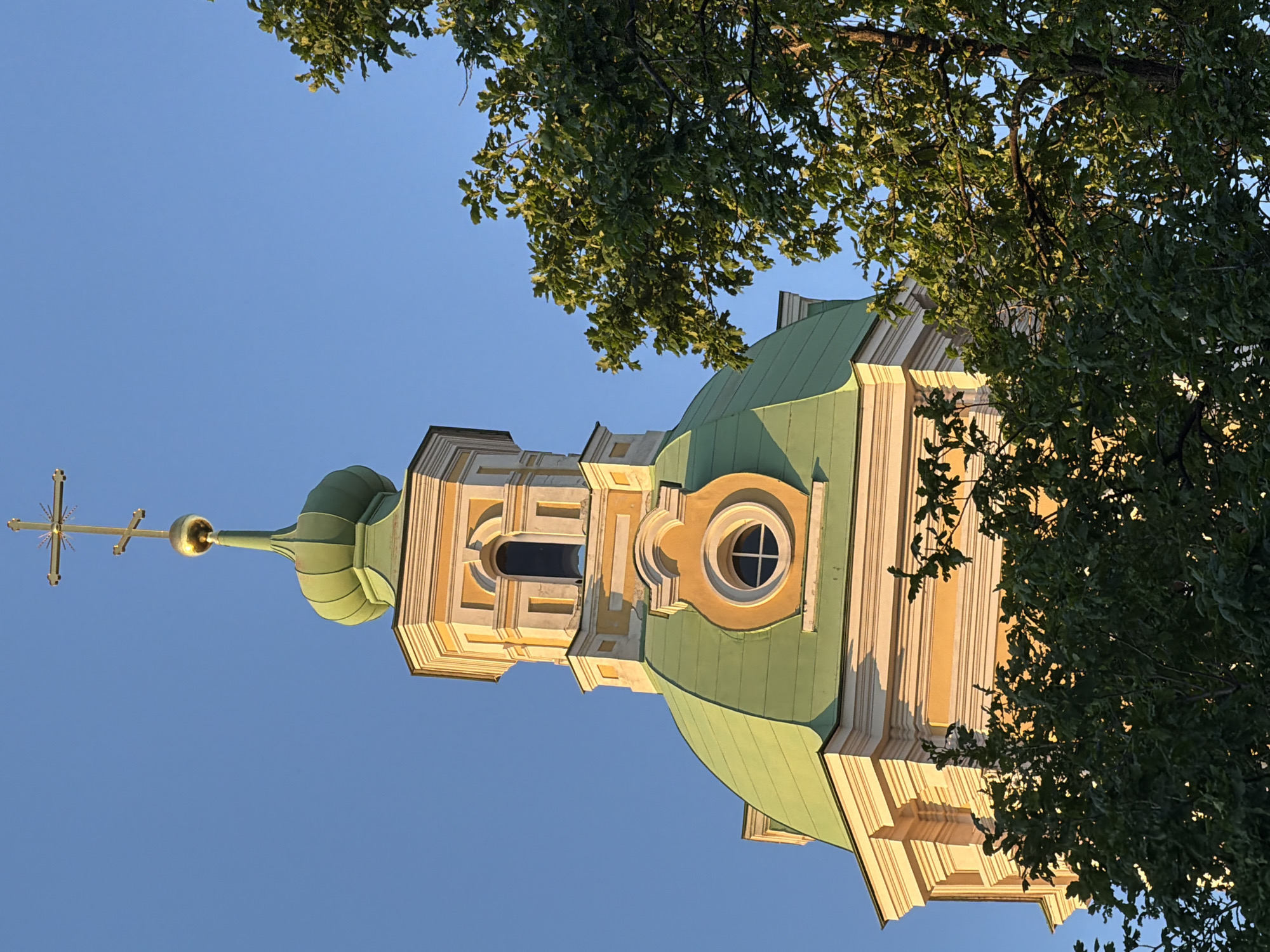The image displays the upper sections of the Vladimirskaya Church (Church of the Vladimir Icon of the Mother of God) in Saint Petersburg, Russia. The structure features a prominent light green dome as the visible base, transitioning into a pale yellow multi-sided tier with white architectural detailing. An oculus window, framed in white, is centered on the yellow facade above the dome. Above this, a segment of a tall, arched window with a dark interior is visible on the multi-sided tier. The structure culminates in a smaller, ribbed light green cupola, topped with a golden finial resembling a small onion dome. This finial is surmounted by a golden Russian Orthodox cross, characterized by three horizontal bars (the lowest slanted) and radiating lines behind the uppermost bar. The building's facade is illuminated by direct sunlight, casting shadows on the architectural elements and highlighting the pale yellow and white surfaces. A clear, vibrant blue sky forms the background. The lower right and parts of the left side of the frame are partially obscured by dense green foliage from deciduous trees.