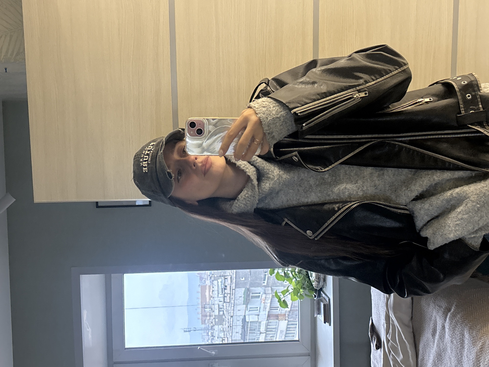 A young woman is taking a selfie indoors, with a cityscape visible through a window. She wears a grey sweater, a black leather jacket, and a dark-colored baseball cap with visible text. The phone she's using appears to be an iPhone. The background shows tall apartment buildings, consistent with an urban residential area in Krasnoyarsk, Russia, based on the architectural style of the buildings visible through the window. A potted plant is also on the windowsill. The interior background consists of a light-colored wall.