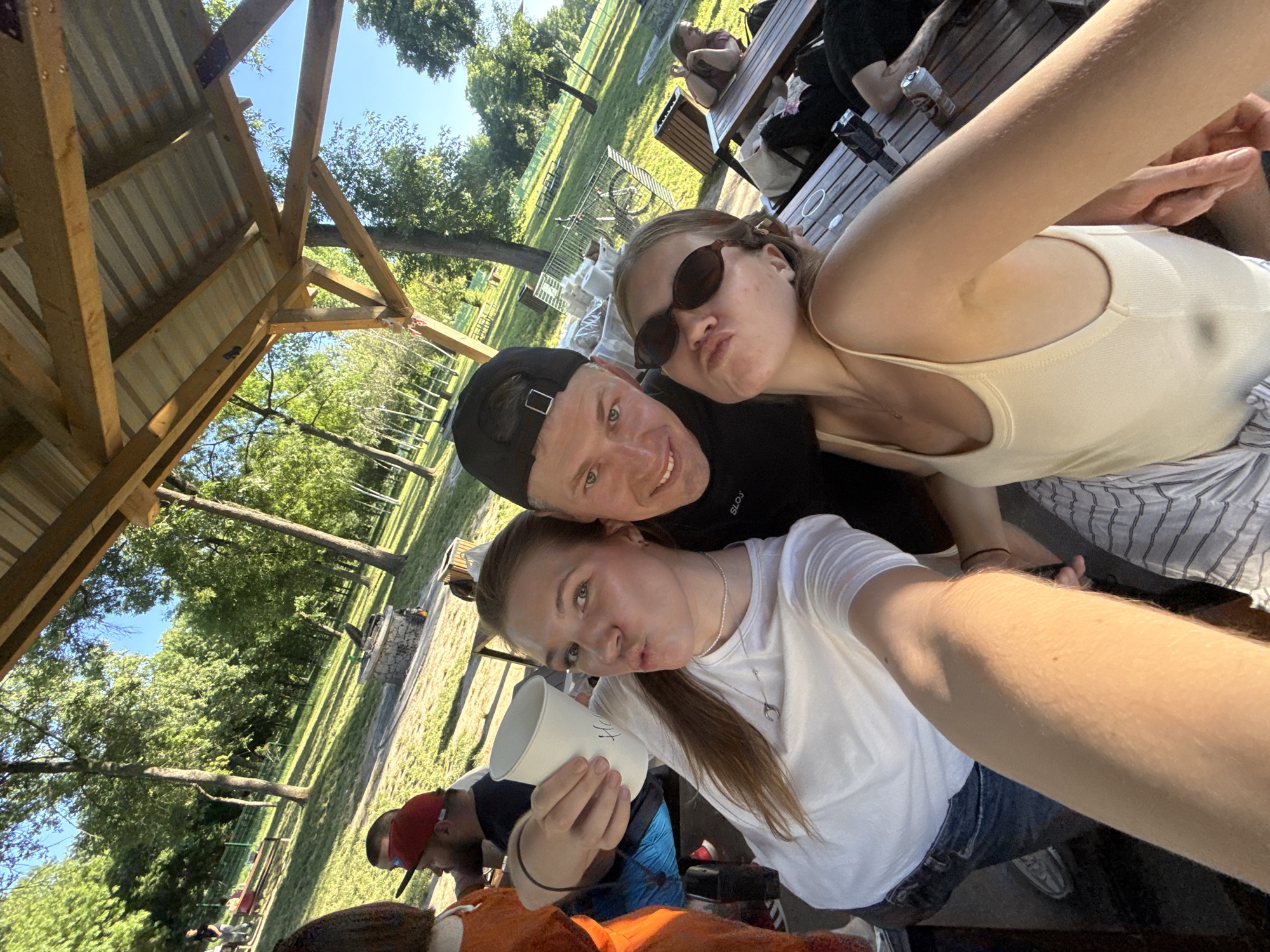 The photograph captures a group of three individuals in a seemingly casual setting, likely outdoors. The photo is taken at an angle, as if someone is holding the camera at arm's length for a selfie.
A young woman, holding the camera, is dressed in a white t-shirt and denim jeans. She has her hair pulled back in a ponytail and is holding a white cup. To her right, a man is wearing a black cap and a black t-shirt. He is smiling at the camera. Next to him is a young woman wearing sunglasses and a light-colored top.
The background suggests an outdoor environment with trees and a grassy area. In the distance, other people can be seen sitting at picnic tables. There's a structure resembling a shelter or gazebo visible on the left side of the frame.
Based on the information provided, the location is in the city of Cukarica Urban Municipality, Serbia. Without more contextual clues, the exact location in the municipality is difficult to determine. The presence of the picnic tables suggests it could be a park, but further information would be needed to confirm.
