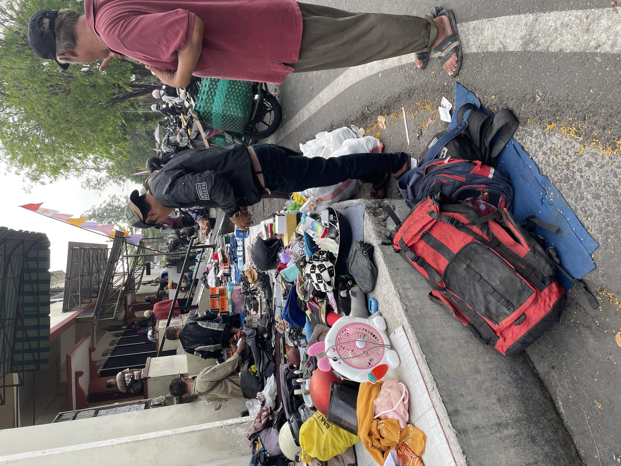 Two men are standing on a sidewalk next to a street. One man, wearing a red polo shirt and olive green pants, is gesturing with his right hand and looking towards the left. The other man, wearing a black jacket with white accents and dark pants, is also looking towards the left and appears to be examining items on display.
The sidewalk is covered with various objects, including clothing, bags, a pink fan shaped like a rabbit, helmets, and other miscellaneous items. Some items are spread out on the ground, while others are arranged on a tiered concrete structure. Bags, including a large red and black duffel bag and a blue backpack, are also present on the ground.
In the background, there are buildings, trees, and parked motorcycles. Flags are visible, and it appears to be an outdoor market or street vendor scene. The ground is asphalt with white lane markings. The overall environment suggests an urban outdoor setting.