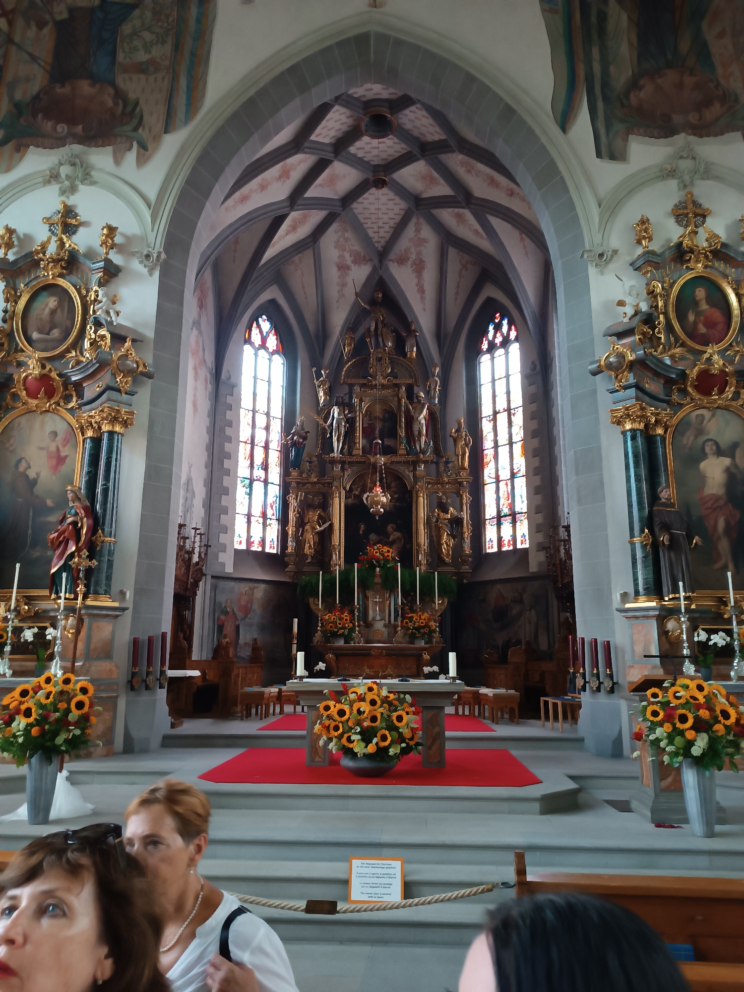 The image captures the interior of a church, likely the St. Mauritius Catholic Church in Appenzell, Switzerland, given its ornate Gothic architecture. The focal point is the altar, adorned with a large, golden altarpiece featuring multiple statues and intricate carvings. Stained-glass windows illuminate the space, casting colorful light on the stone walls and the vaulted ceiling, which is decorated with frescoes. Two large vases filled with sunflowers and other flowers flank the altar. In the foreground, several people are visible, though their faces are partially obscured. One woman in the lower left is looking upwards and to the left, her expression unreadable. Another woman, to her right, is wearing a white top and a pearl necklace, looking towards the altar. The scene is well-lit, highlighting the rich details of the church's interior.