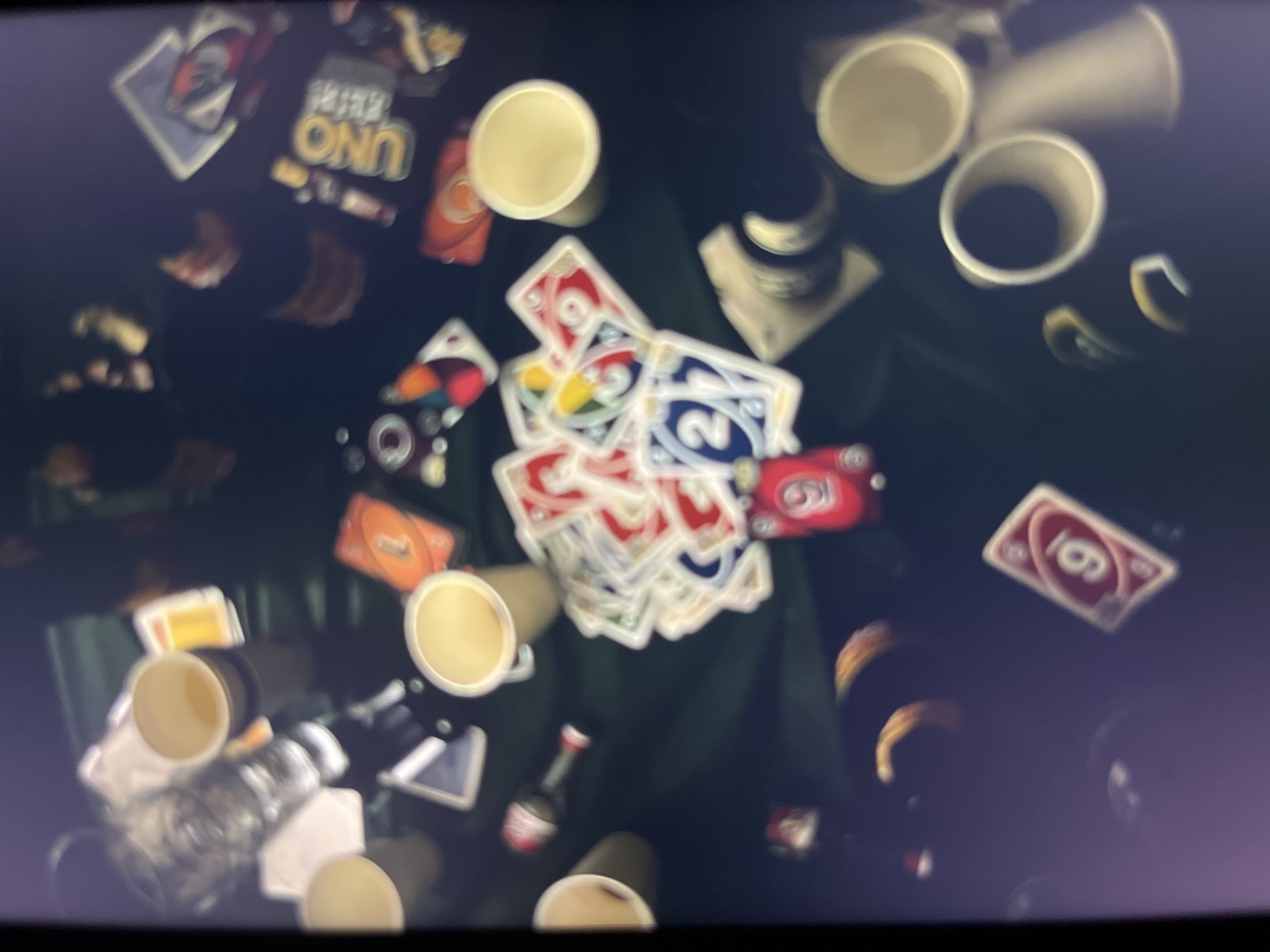 The image displays a dark, cluttered tabletop scene, possibly a gathering or party, with several UNO playing cards scattered across a dark green surface. Several paper cups are visible, some empty and some containing a light-colored liquid. There are also some blurred bottle-like objects, possibly drinks. In the foreground, a red UNO card with the number 9 is prominently displayed, along with other UNO cards showing numbers and colors like red, blue, and yellow. The lighting is dim, and the overall image has a somewhat blurry quality, suggesting it was taken with a handheld camera in low light conditions. There is no discernible indication of Bukuru, Nigeria, or any specific location within it. The scene appears to be an indoor setting, with the focus on the game and related items.