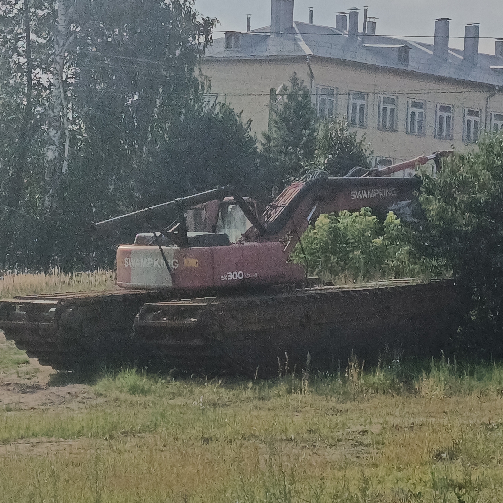 An amphibious excavator, identified by the text "SWAMPKING" and "SK300" on its body, is situated on a muddy platform. The excavator is a large, orange and red machine with a boom and bucket. It is surrounded by green foliage and trees. In the background, a multi-story building with several windows and chimneys is visible. The scene appears to be in an outdoor, possibly industrial or construction site setting, within the city of Kambarka, Russia, judging by the architectural style of the building. The ground is covered with grass and some dirt patches. There are no people visible in the image.