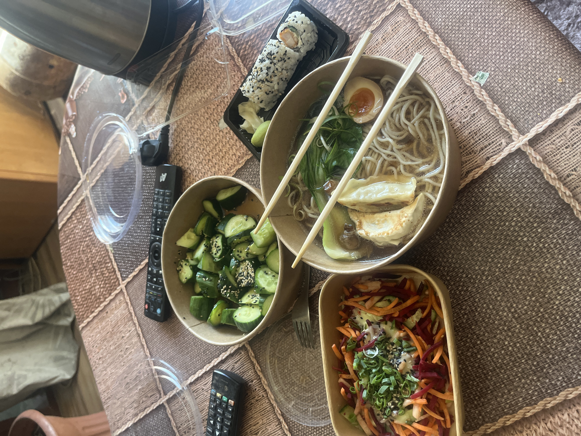 The image shows a table set with an assortment of takeout food. There is a bowl of ramen with noodles, broth, egg, gyoza, and greens; a container of cucumber salad with sesame seeds; a container with shredded carrots and beets topped with green onions and sesame seeds; and a tray of sushi with a side of ginger and wasabi. Chopsticks rest on top of the ramen bowl. Two remote controls and clear plastic containers are also visible on the table. The table has a brown woven placemat. The location is unspecified, but since the request specifies Sofia, Bulgaria, it may be assumed that the location is a residence or restaurant in that city. There are no people visible.
