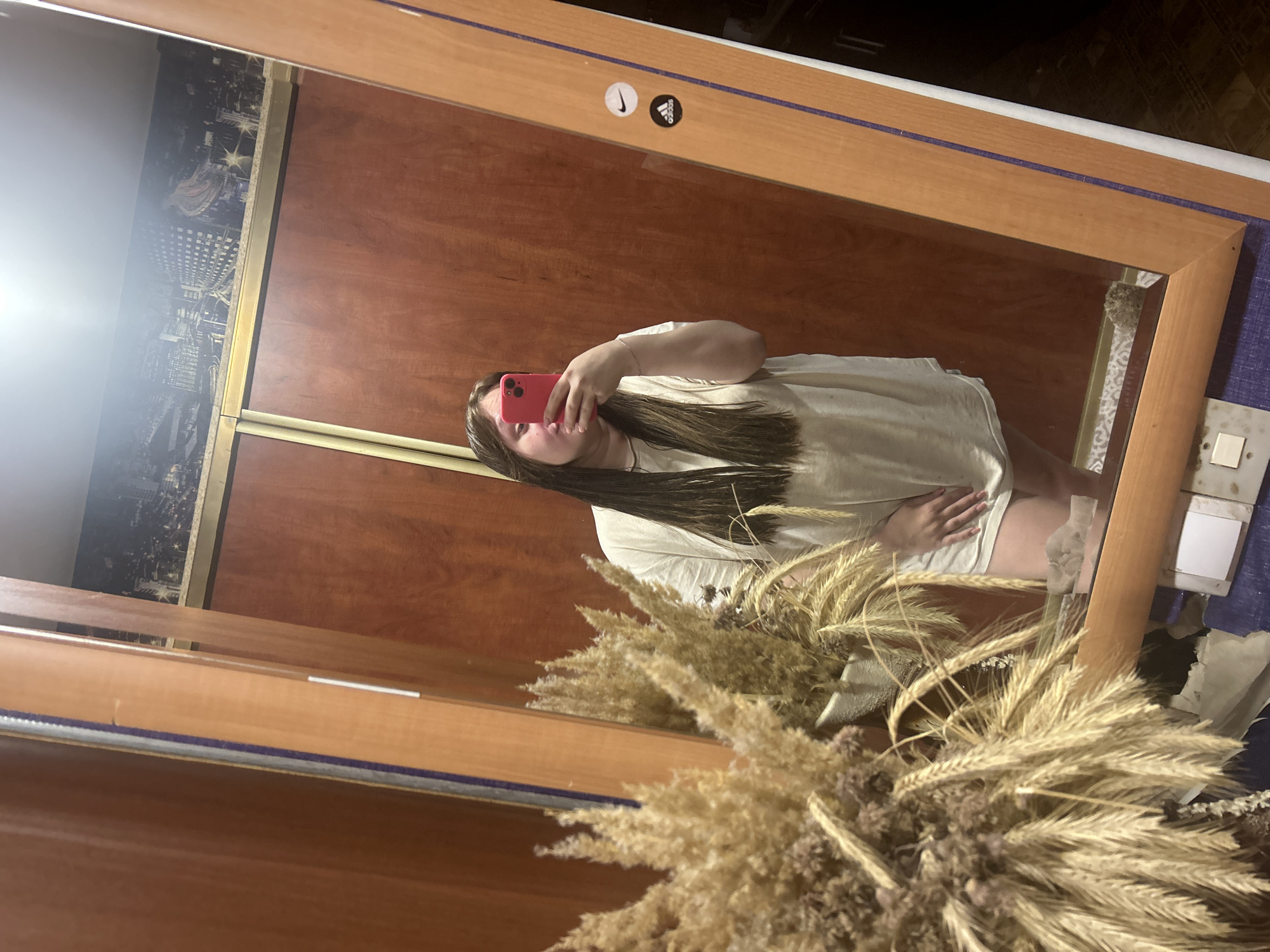 A woman is taking a selfie in a mirror. The mirror is in a room with wooden walls and a decorative strip showing a cityscape, possibly of Ольховка, Russia. The mirror has a wooden frame and two stickers, one with a Nike logo and another with an Adidas logo. There is a bouquet of dried wheat and other dried plants in the foreground obscuring the view of the woman. The woman is holding a red phone in her left hand. The woman is wearing a white t-shirt.
