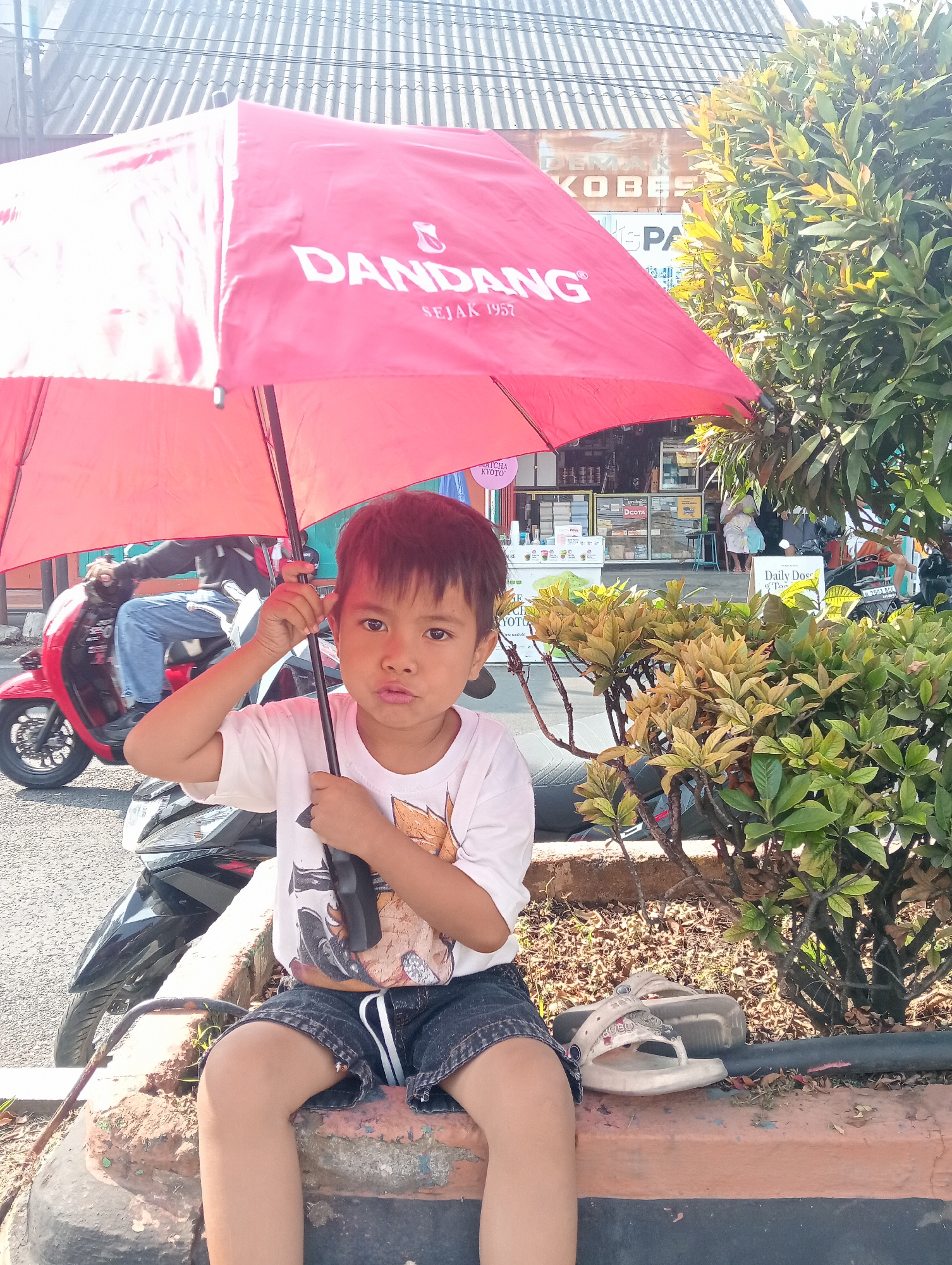 A young boy wearing a white t-shirt with a graphic print and denim shorts sits on a concrete ledge. He is holding a red umbrella open above him. A red motorcycle is parked behind him, and in the background, there are storefronts and a person riding a motorcycle. The scene appears to be on a street in Bintoro, Indonesia, with a building and trees visible. The lighting suggests it is daytime.