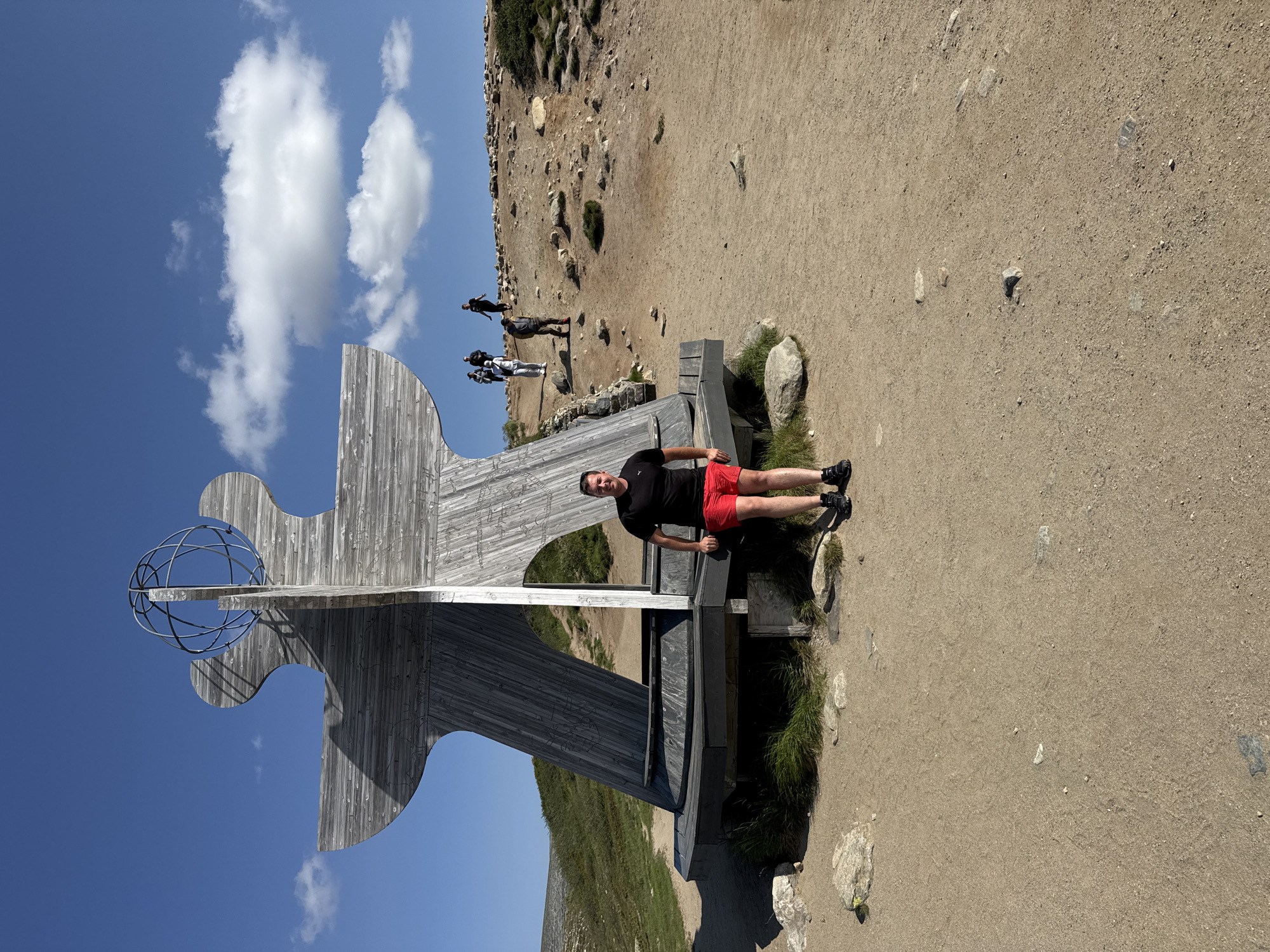A man stands in front of a large wooden sculpture with a metal globe on top. The sculpture is abstract and appears to be made of weathered wood planks. The man is wearing a black t-shirt and red shorts. He is smiling and leaning against the base of the sculpture. Behind him, several other people are walking up a rocky, dirt path on a hillside. The environment is a natural landscape with sparse vegetation and a clear blue sky with some white clouds. The location is the Arctic Circle monument in Storforshei, Norway.