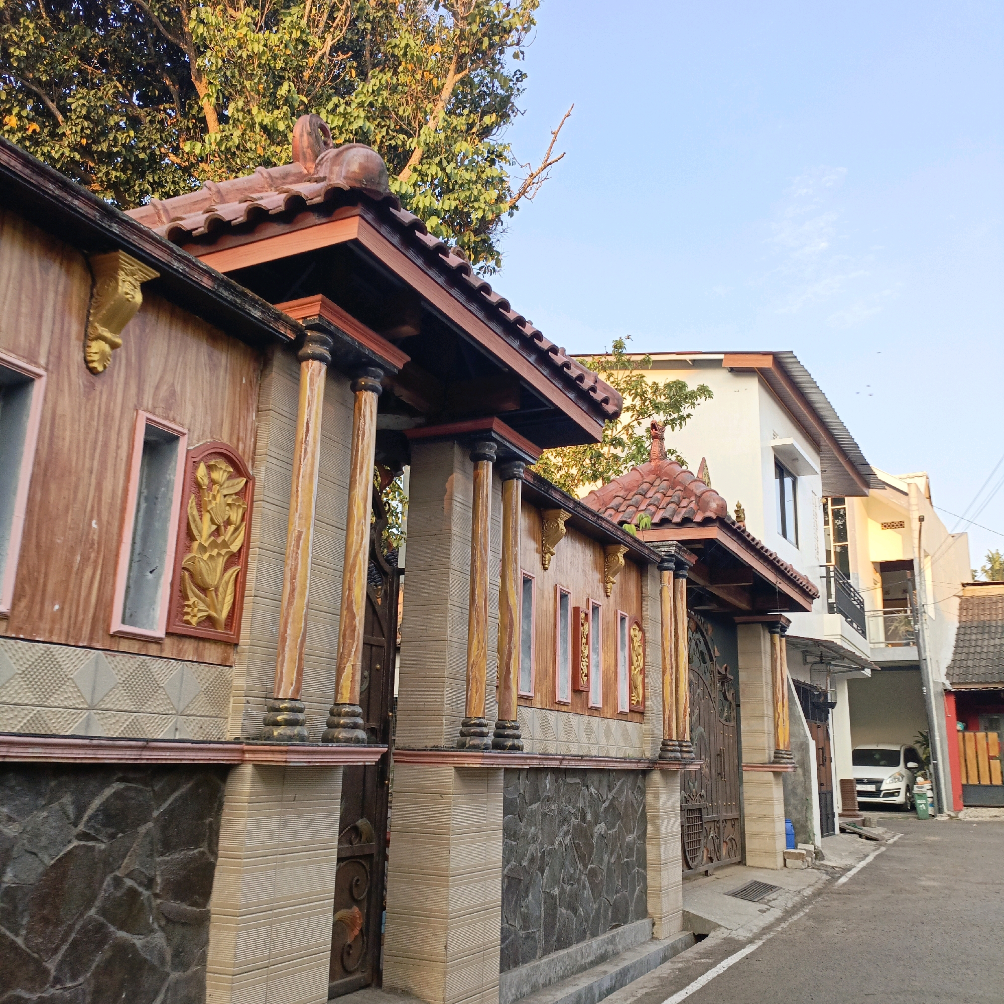 The image depicts a street scene in Salatiga, Indonesia. In the foreground, a decorative wall with tiled pillars and a red tiled roof separates a property from the street. The wall features a faux wood-grain pattern, stone cladding at the base, and ornate golden floral reliefs. To the right, a paved street runs alongside the wall. Further down the street, a white car is parked under a carport attached to a white multi-story building. The background shows more residential buildings and a large tree with green foliage. The sky is clear and blue.