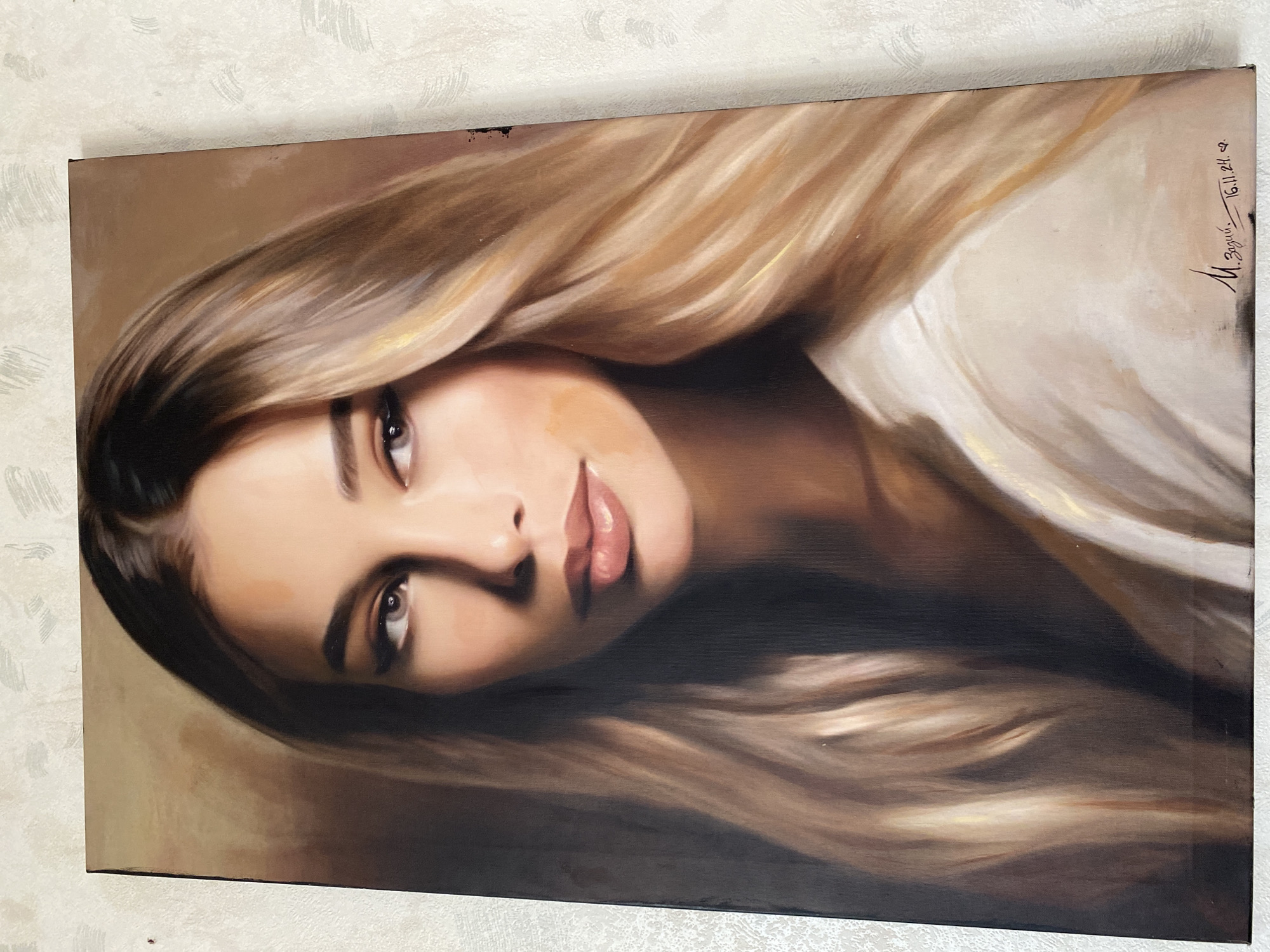A painting of a woman with long, blonde hair is hanging on a wall. The painting is a close-up portrait of the woman's face, with her hair flowing around her. The woman has light skin and brown eyes. The signature on the painting reads "M. Zagid...." and "16.11.24." The painting is located on a white wall, which may be in a house or apartment. There is no evidence the image is taken in the city of Chaikovsky, Russia.
