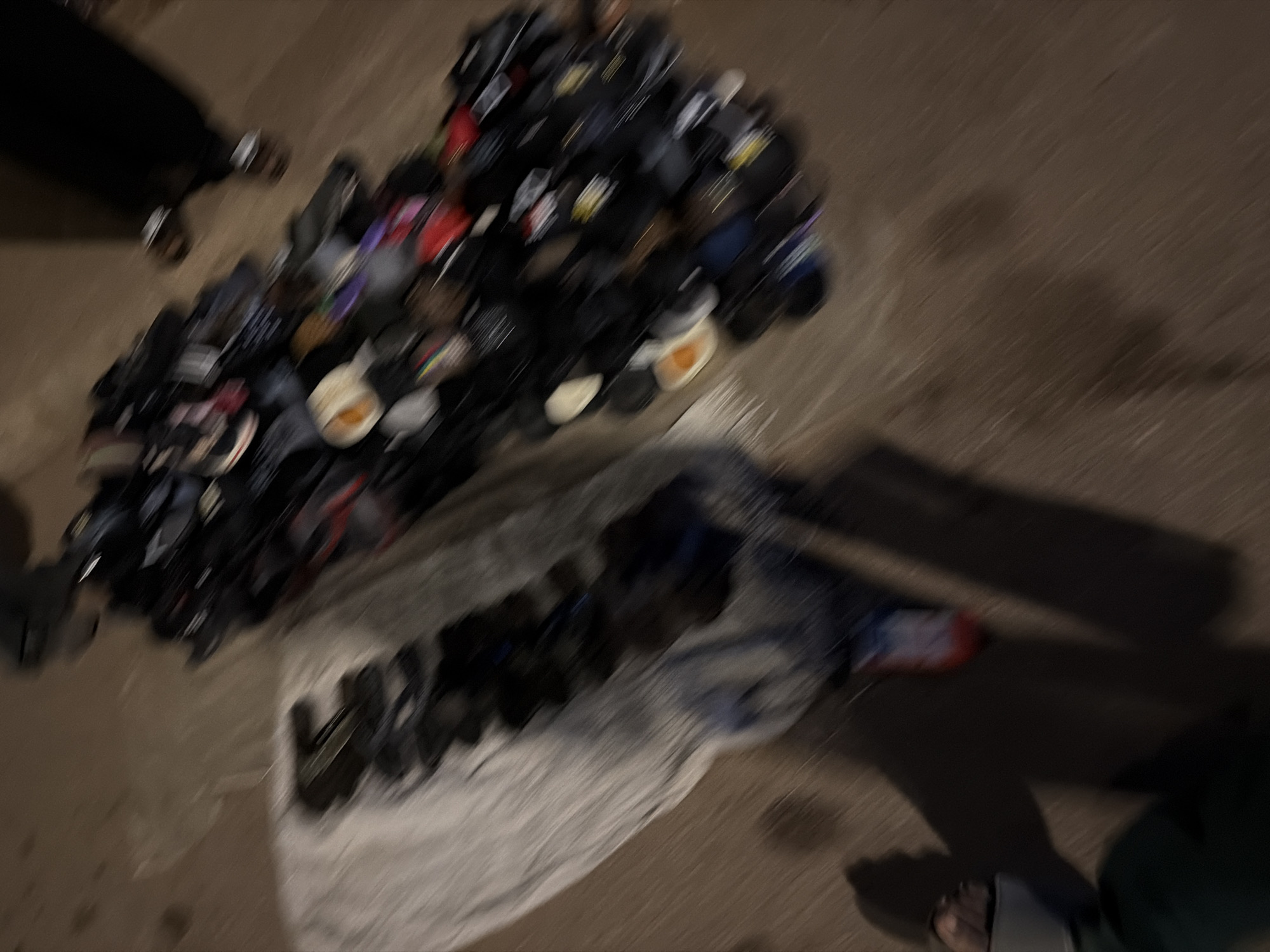 The image depicts a busy outdoor market scene in Bukuru, Nigeria, possibly a street stall. In the foreground, a person's legs and feet are visible, wearing sandals, and partially shadowed by darkness. To the left, a pile of various footwear, including sandals and shoes, is displayed on a light-colored cloth. To the right, a larger, more densely packed collection of shoes and sandals is arranged on the ground, some appearing to be men's and women's footwear, with various colors and designs visible. The ground is a paved or concrete surface, common in urban areas of Nigeria. The overall impression is one of a bustling commercial environment, likely during evening hours given the lighting conditions and shadows.