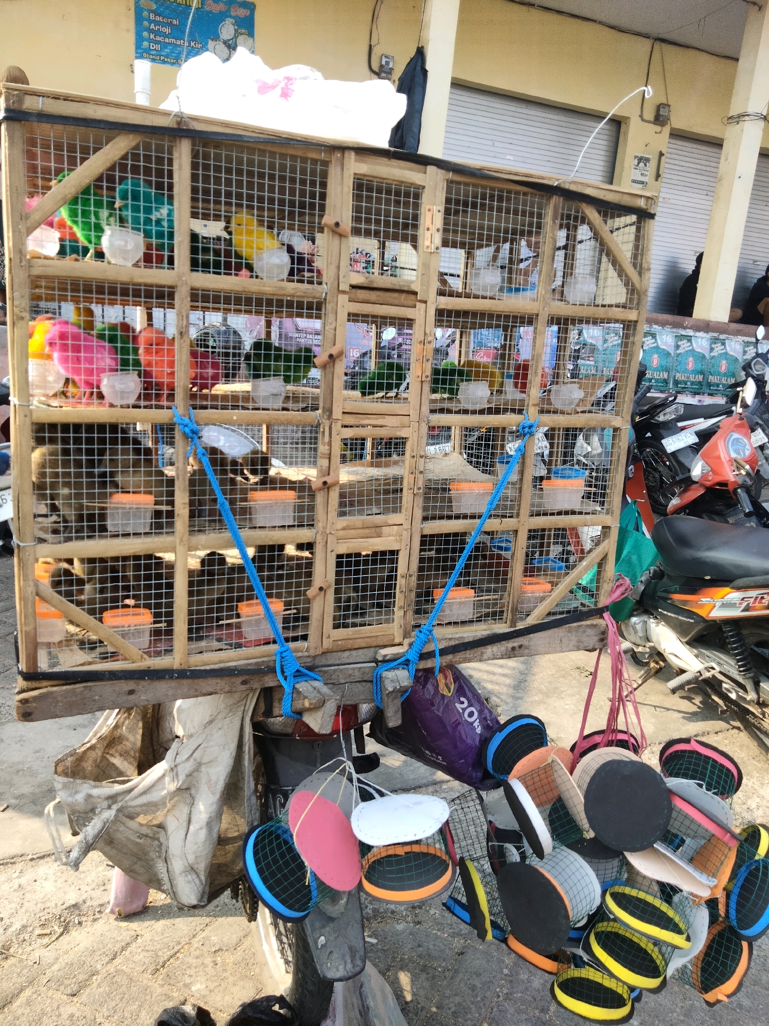 In Sawahan, Indonesia, a cart, likely attached to a motorcycle, is laden with a wooden bird cage. The cage contains several colorful birds, possibly parrots or parakeets, in various shades of green, yellow, and pink. Orange plastic food and water containers are visible inside the cages. Below the cage, a collection of colorful, netted hats or baskets are displayed, some hanging from the cart. In the background, a building with roll-up metal doors suggests a market or shop setting. Motorcycles are parked nearby, indicating a public area. The ground is paved with concrete and dirt.