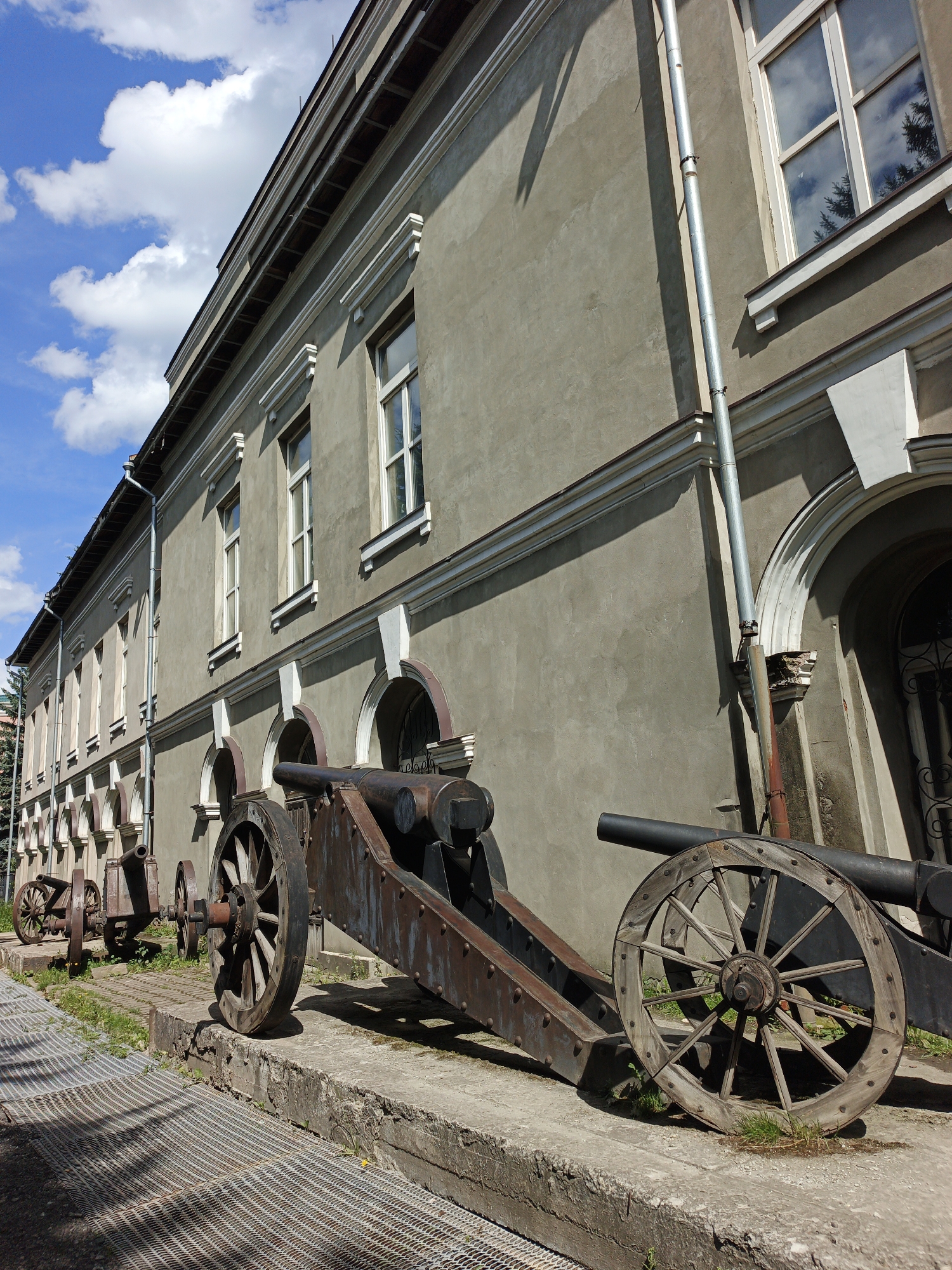 This is a low-angle, full shot photograph taken outdoors of the exterior of a long, gray building with a blue sky and scattered white clouds in the background. The building has multiple windows with white trim, and an arched arcade runs along the lower portion of its facade. In the foreground, two antique cannons are positioned on a concrete ledge in front of the building. The cannon on the right is larger and more prominently featured, with a thick black barrel and a large wooden spoked wheel. The cannon on the left is smaller, appearing older and more weathered, with a narrower barrel and smaller wooden spoked wheels. Both cannons are made of dark metal and show signs of age and rust. A metal grate walkway is visible in the lower left corner of the image. The scene appears to be a historical site or museum exhibit in the city of Zlatoust, Russia. There are no people or animals present in the image.