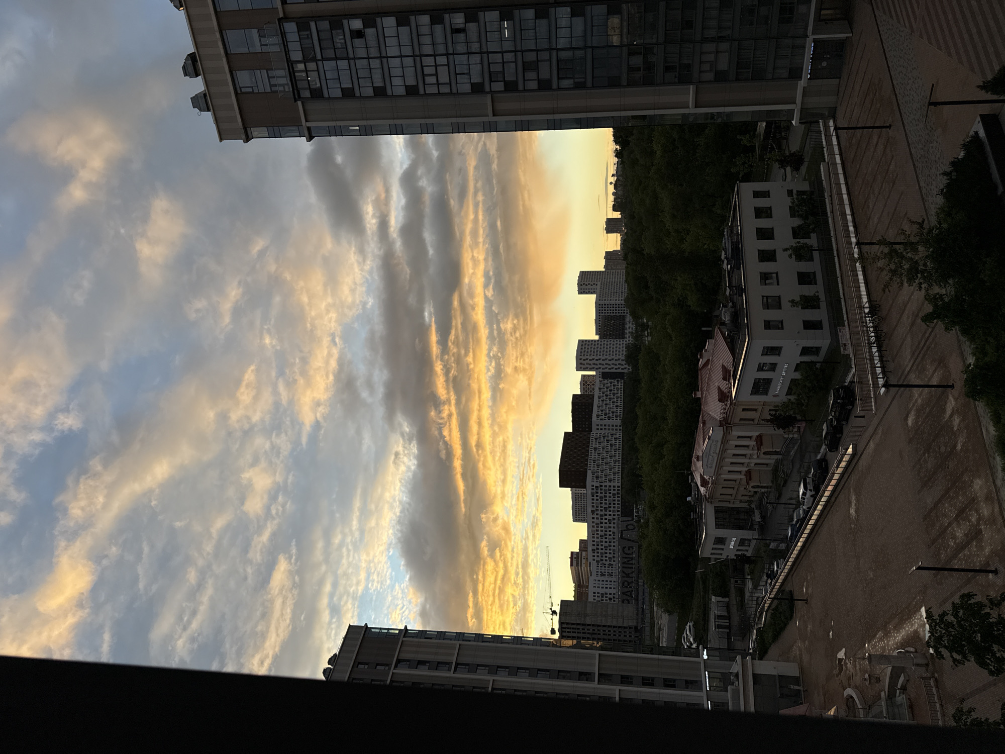 The image depicts an urban cityscape at dusk. Tall buildings flank a central view of a sky filled with striated clouds, illuminated with orange and yellow hues from the setting sun. A line of trees is visible further in the distance, interspersed with more buildings. Below the trees and buildings, there is a street with parked cars and pedestrian walkways. The photograph is likely taken from a high vantage point within one of the taller buildings. The location is Khimki, Russia, based on the style of buildings and urban planning. A crane is visible in the distance. A building has "PARKING" written on the side.