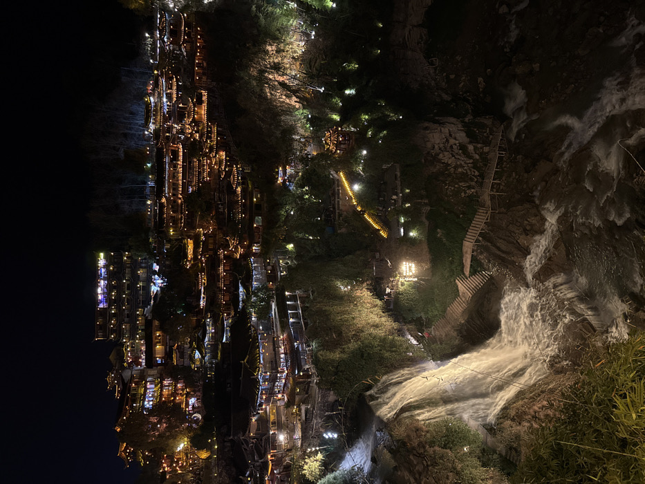 This image presents a nighttime panoramic view of Furong Ancient Town, China, centered on its illuminated hillside architecture and a prominent waterfall. Numerous multi-tiered buildings, constructed in traditional Chinese architectural styles with dark, curving roofs and wooden elements, are densely arrayed across a steep hillside in the midground and background. These structures are extensively lit with warm yellow and orange lights, creating a glowing urban landscape against the dark sky.
In the foreground and lower midground, a large, powerful waterfall cascades down a rocky cliff face. The rushing water is brightly illuminated by white artificial lights, highlighting its foamy descent into a river or stream that flows over smaller rapids at the bottom of the frame. The terrain surrounding the waterfall and town consists of rugged cliffs and hillsides covered with dark green vegetation, including bamboo and trees.
Several wooden walkways and staircases, strung with warm yellow lights, are visible winding along the cliff edges, particularly on the right side, leading down towards the base of the waterfall and the river. No human figures or specific activities are discernibly visible within the frame. The scene is dominated by artificial light sources, creating a luminous and dramatic nocturnal environment.