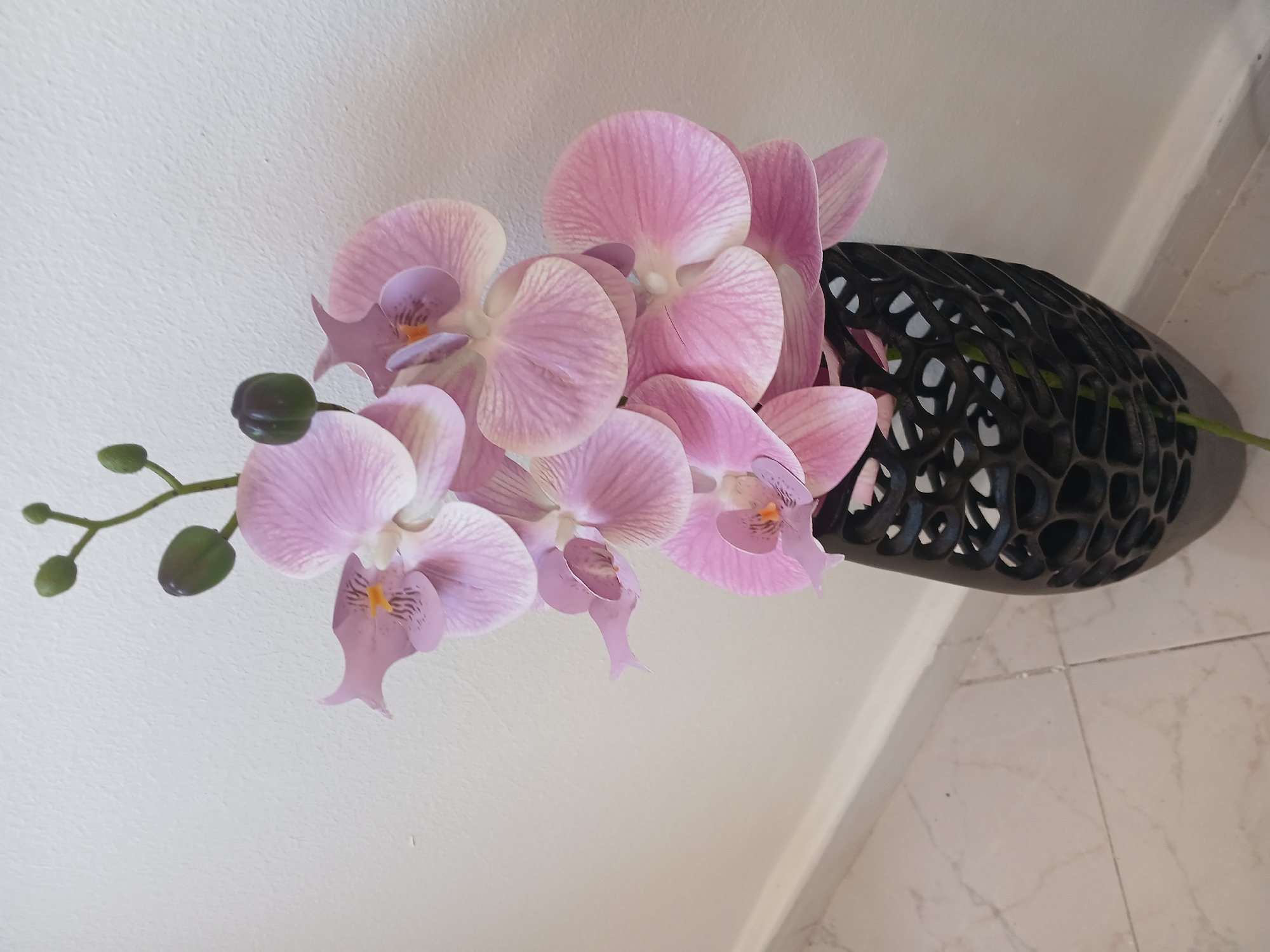 The image shows a close-up, slightly angled view of artificial pink orchids arranged in a dark, geometrically patterned vase. The orchids are in full bloom, with detailed petals displaying pink and light purple hues. A branch with green buds extends to the left. The vase is dark and appears to be made of a material with a lattice-like structure. The background is a plain white wall, with a section of light-colored tiled floor visible in the bottom right. The location is described as Unknown, Unknown.