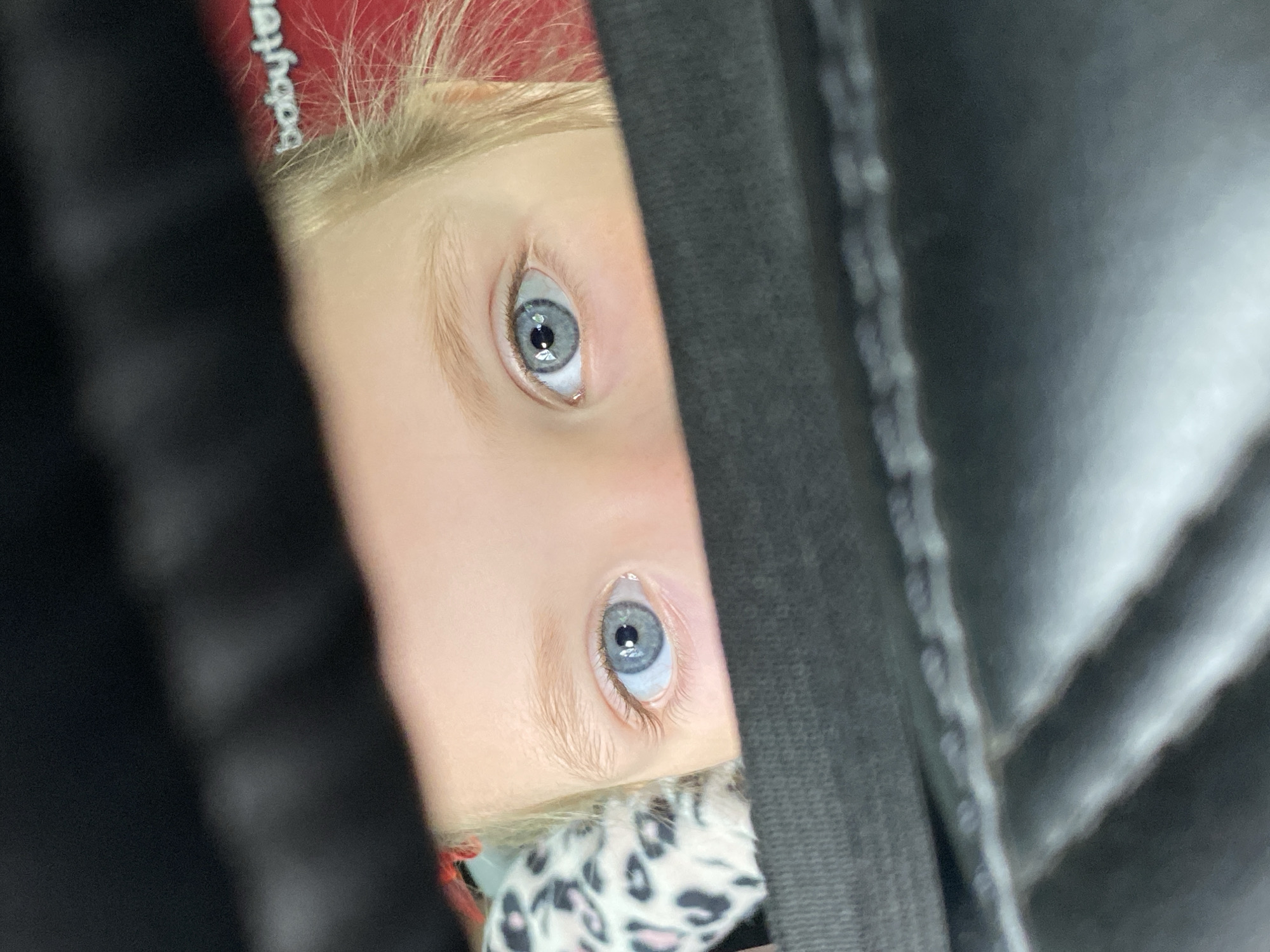 A close-up, upside-down shot shows a young child's face partially obscured by a dark, hooded garment. The child has light blonde hair and blue eyes, which are the focal point of the image. Part of the child's cheek and forehead are visible, along with a glimpse of a patterned cloth. The hood appears to be black and made of a dense material with a visible seam. There is no information in the image to suggest this was taken in the city of Votkinsk, Russia.