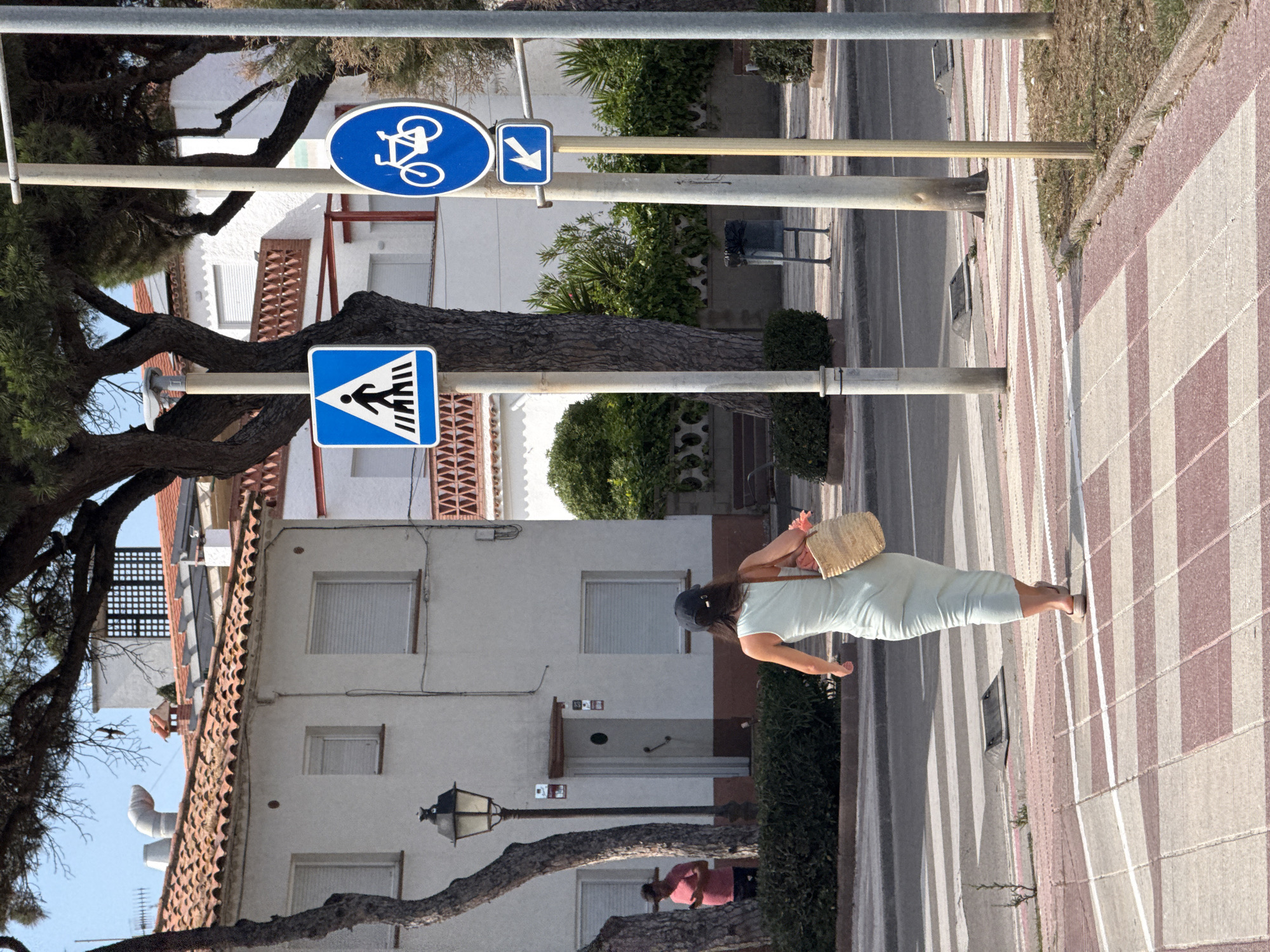 A woman wearing a light-colored, form-fitting dress and a dark baseball cap walks across a crosswalk, carrying a straw bag over her right shoulder. She is seen from behind, moving from left to right in the frame. In the background, a white building with orange-tiled roofs is visible, featuring windows with closed shutters and decorative brickwork. A lamppost stands near the building. To the right of the woman, two street signs are mounted on poles: a blue square sign with a white pedestrian symbol indicating a crosswalk, and below it, another blue sign with a white bicycle symbol and an arrow pointing left. The area is paved with a checkered pattern of light and dark pinkish tiles. Trees with dark bark and green foliage are present on the left and right sides of the scene. The sky is clear and blue.