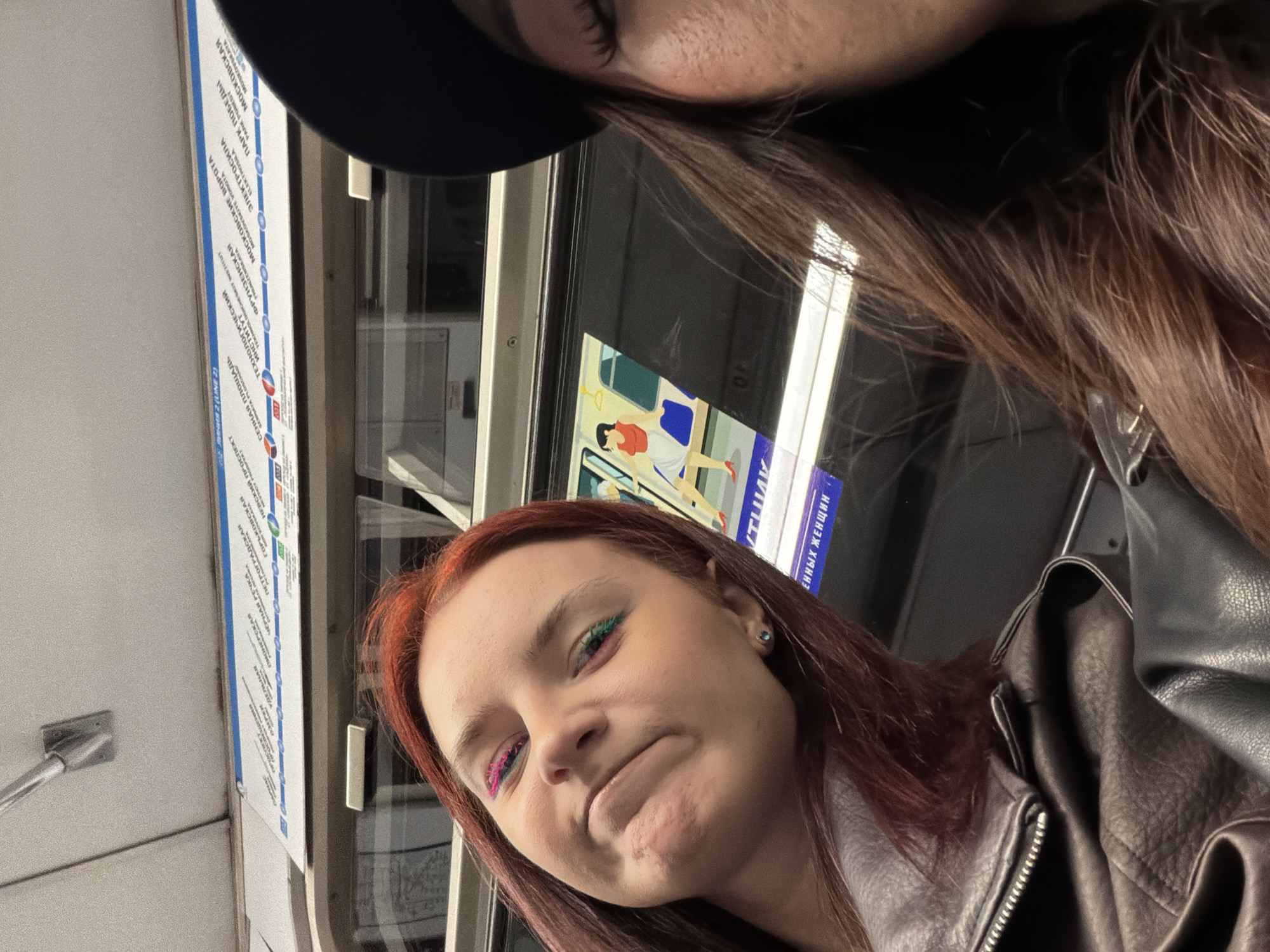 Two young women are inside a subway car, likely in a city. The woman on the left has dark red hair and is wearing a grey leather jacket. She is looking towards the camera with a slightly squished expression and colorful eyeshadow on her eyelids. The woman on the right is partially visible, wearing a black hat and has long brown hair. Behind them, a subway map is displayed on the wall, showing different lines and stations. There is also a graphic on the window of a person standing by a subway door. The overall lighting suggests an indoor environment, typical of public transportation.