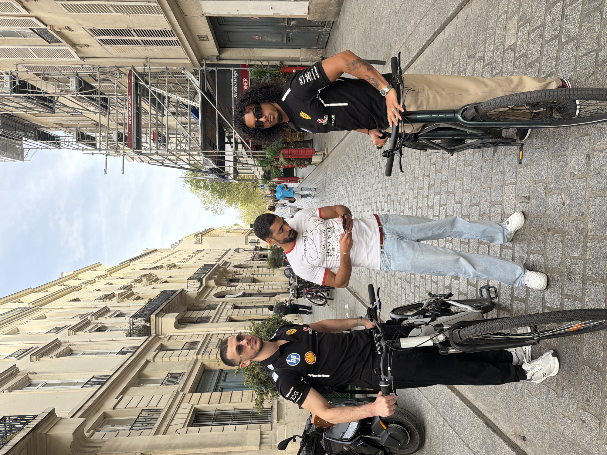Three men stand on a cobblestone street in Paris, France, flanked by tall buildings. Two of the men are on bicycles, one wearing a black polo shirt with corporate logos and black pants, and the other wearing a similar shirt with khaki pants and sunglasses. The third man stands between them in a white t-shirt and light blue jeans, looking down at his phone. Scaffolding is visible on the buildings. The surroundings include parked bicycles and pedestrians in the background.