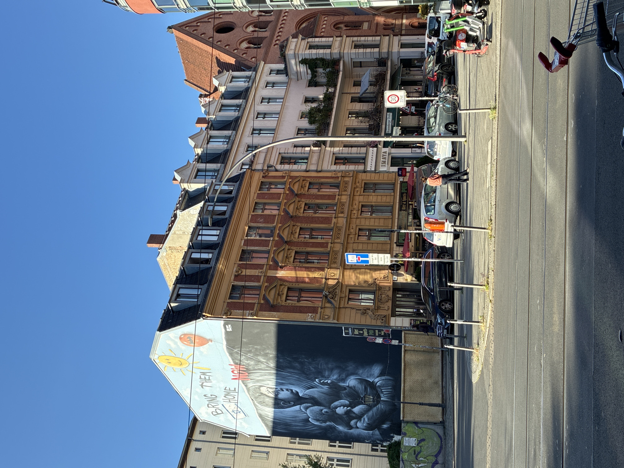 The image captures a sunny day in Berlin, Germany, showcasing a street scene with various buildings and a prominent mural. On the left, a large mural adorns the side of a building, depicting a child holding a teddy bear, with a sun and balloons in the sky. The text "BRING THEM HOME NOW" is painted above the child, along with a Star of David. Adjacent to this mural is a building with a warm, orange facade and intricate architectural details. Further to the right, a lighter-colored building with balconies filled with greenery stands out, followed by a brick building with arched windows, resembling a church. The street in front of these buildings is paved, with tram tracks visible. Several cars are parked and driving on the street. A few people are seen walking or standing near the buildings. Scooters and bicycles are also present in the scene. The sky is clear and blue, indicating good weather conditions. The location appears to be a bustling urban environment, likely in a residential or commercial district of Berlin.