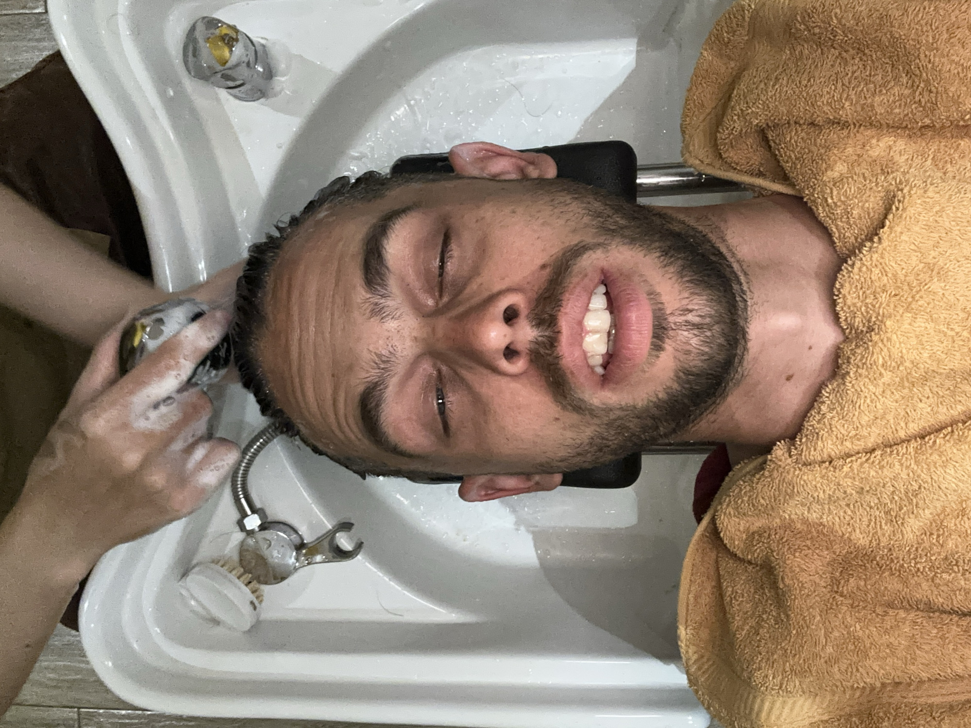 An overhead shot captures a man lying back in a salon wash basin, his eyes closed as his hair is shampooed. A person's hands, covered in foam, are actively massaging the man's scalp. The wash basin itself is white ceramic, with a chrome faucet and a brush visible to the left. The man is draped in a textured, golden-brown towel. Water is visible in the basin and on the man's face and hair. The background appears to be a tiled wall. This scene is taking place in a hair salon in Da Nang, Vietnam.