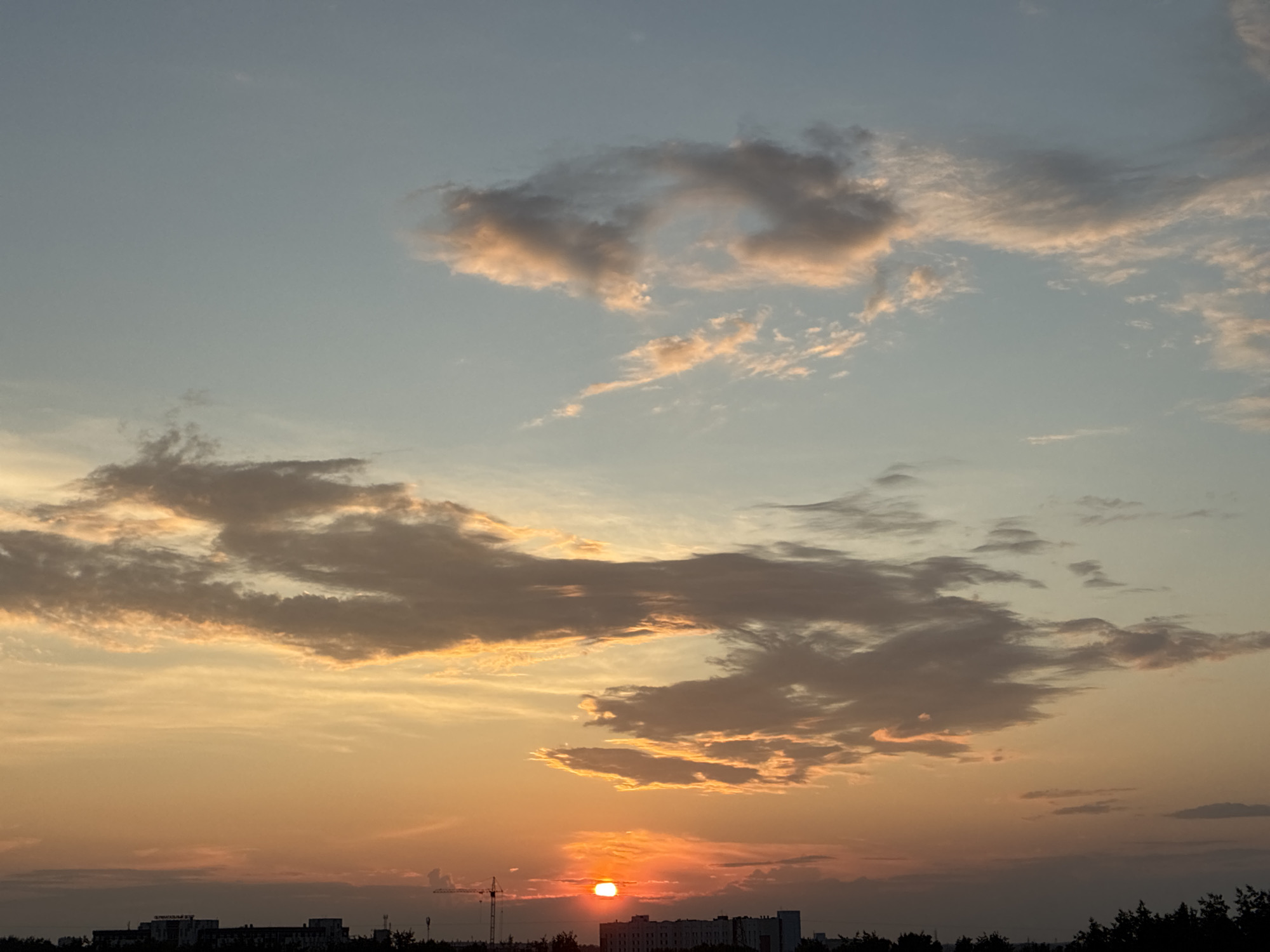 A wide shot captures a sunset over an urban horizon. The sky is a gradient of pale blue at the top, transitioning to warm hues of orange and yellow as it nears the horizon. Scattered clouds are visible, tinged with pink and orange by the setting sun. In the distance, a silhouetted city skyline is present, with buildings and trees forming a dark line against the illuminated sky. A construction crane is also visible on the left side of the frame. The sun itself is a bright, small orb of intense orange, partially obscured by clouds.