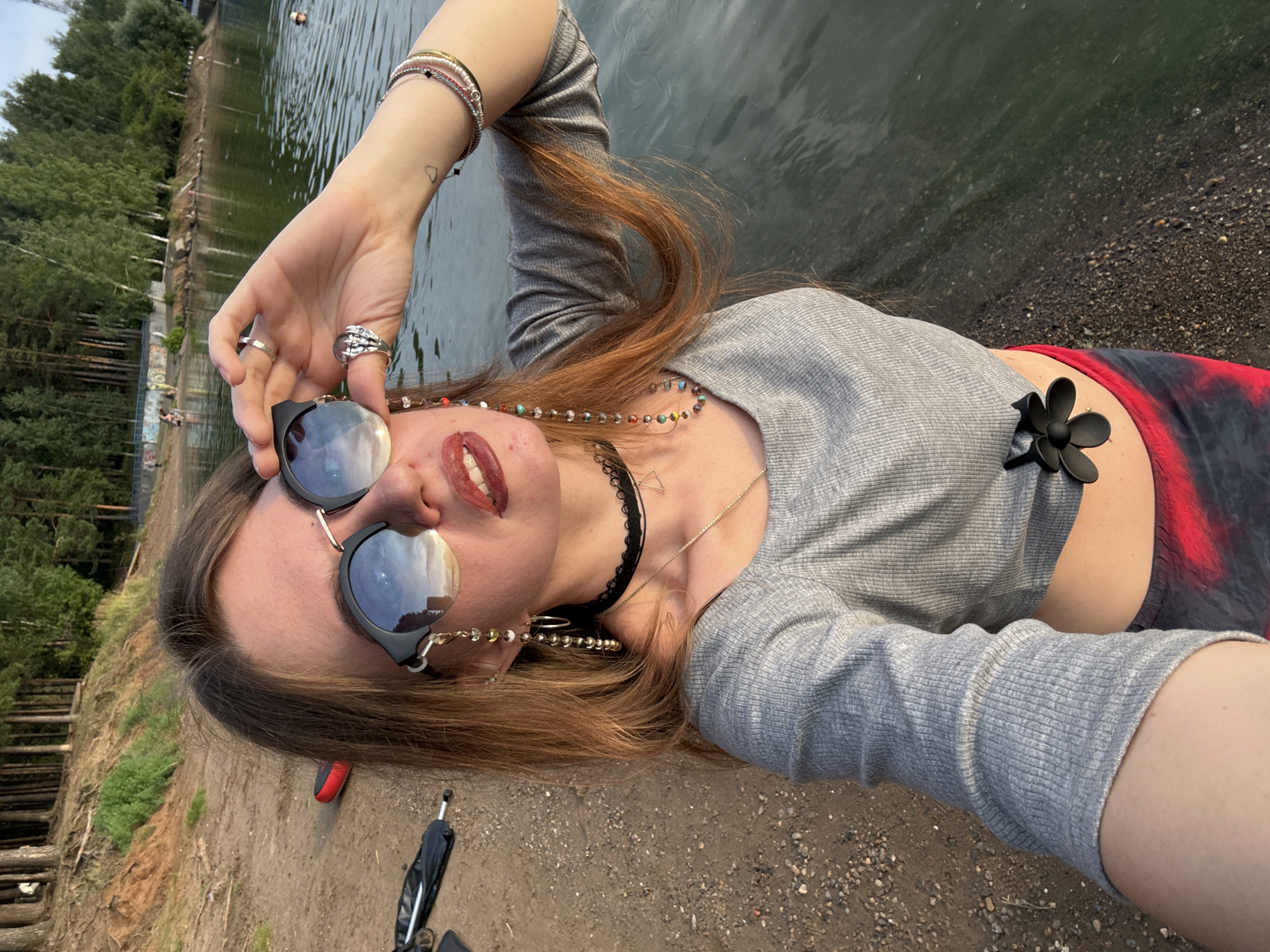 A young woman with long, reddish-brown hair is taking a selfie on a sandy shore next to a body of water. She is wearing sunglasses, a gray crop top, and red and black tie-dye shorts. She is also wearing several rings and bracelets, as well as a black choker necklace with a small triangle pendant. The woman's right hand is holding up her sunglasses, and her left arm is resting by her side. In the background, there are trees and a few other people visible in the water and on the shore. The lighting suggests it is late afternoon or early evening.