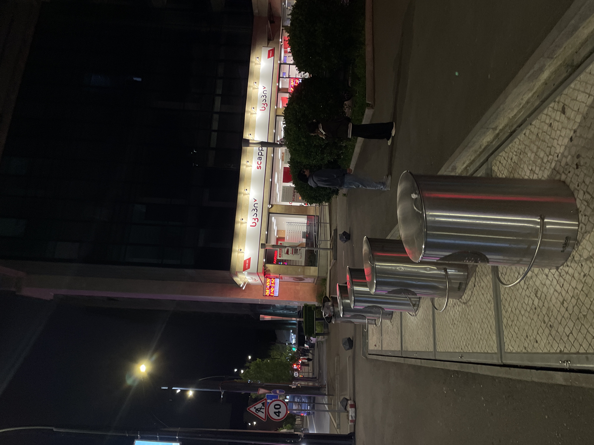 The image is taken at night in Tbilisi, Georgia, showing a street with a Scappino store in the background. Two people stand near the store. Several cylindrical metal bins with attached lids are arranged along the sidewalk, next to a tiled wall and a metal barrier. Streetlights illuminate the scene, and a street sign indicating a 40 km/h speed limit and pedestrian crossing is visible in the distance.