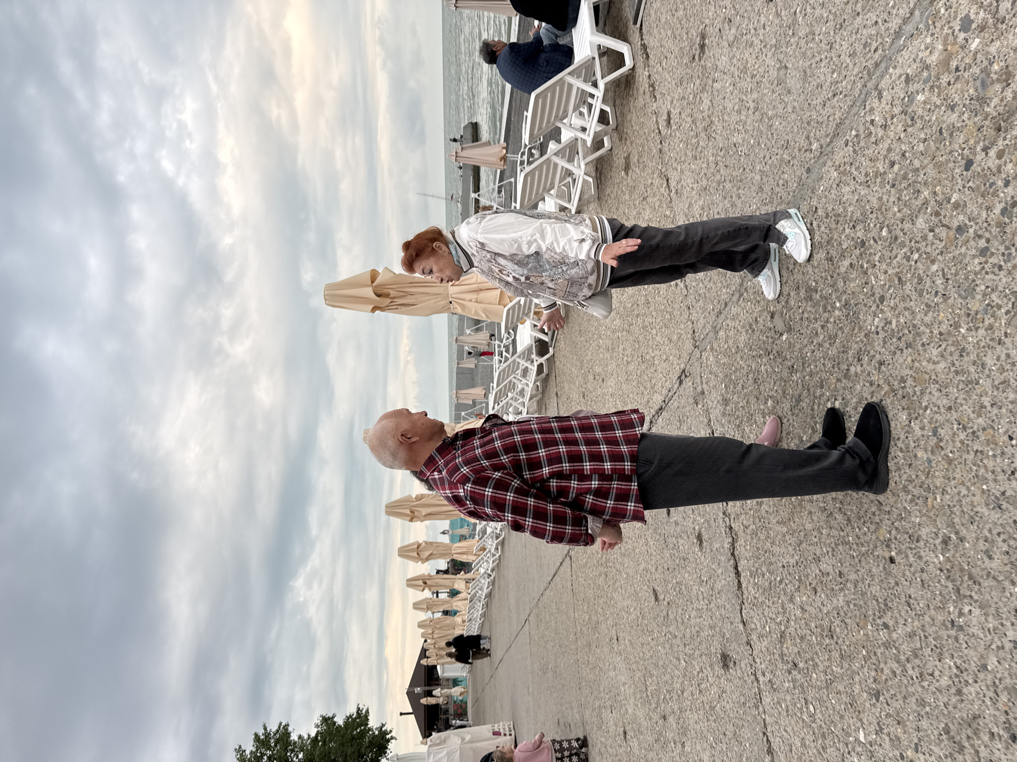 An eye-level photograph shows a man and a woman standing on a concrete walkway near a beach. The image is oriented sideways. The man, on the lower half of the image, is older with a bald head, wearing a red plaid shirt and dark pants. He appears to be looking upward. The woman, on the upper half of the image, has red hair pulled back and is wearing a light-colored patterned jacket and dark pants. She is looking downwards towards the man.
Behind them are rows of closed beige beach umbrellas and white lounge chairs, suggesting a beach scene. Further back, there is a glimpse of the sea and sky, which is cloudy. At the very bottom left corner, there is some green foliage suggesting trees or bushes. The people are on a boardwalk.
The location is assumed to be in Sochi, Russia.