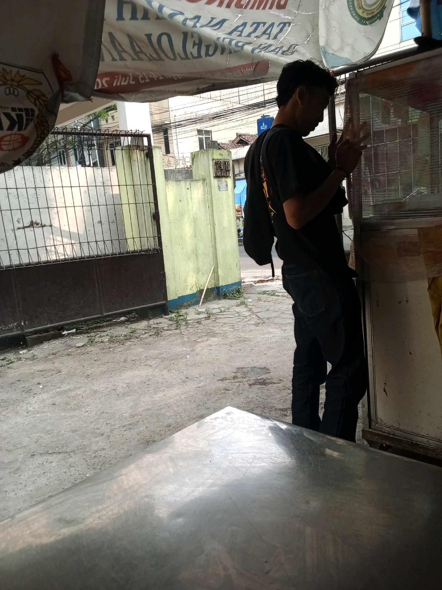 The image depicts a young man in a black t-shirt and blue jeans standing by a stall. He is looking down and appears to be in the process of picking up or adjusting something on the stall's surface. The stall itself is a metal structure with a glass or plastic front, likely used for selling goods. In the background, there's a faded banner with Indonesian writing and a chain-link fence. The environment appears to be an urban setting, possibly a street or marketplace, with a concrete ground and some vegetation. The overall scene is casual and everyday, capturing a moment of the man's interaction with the stall.