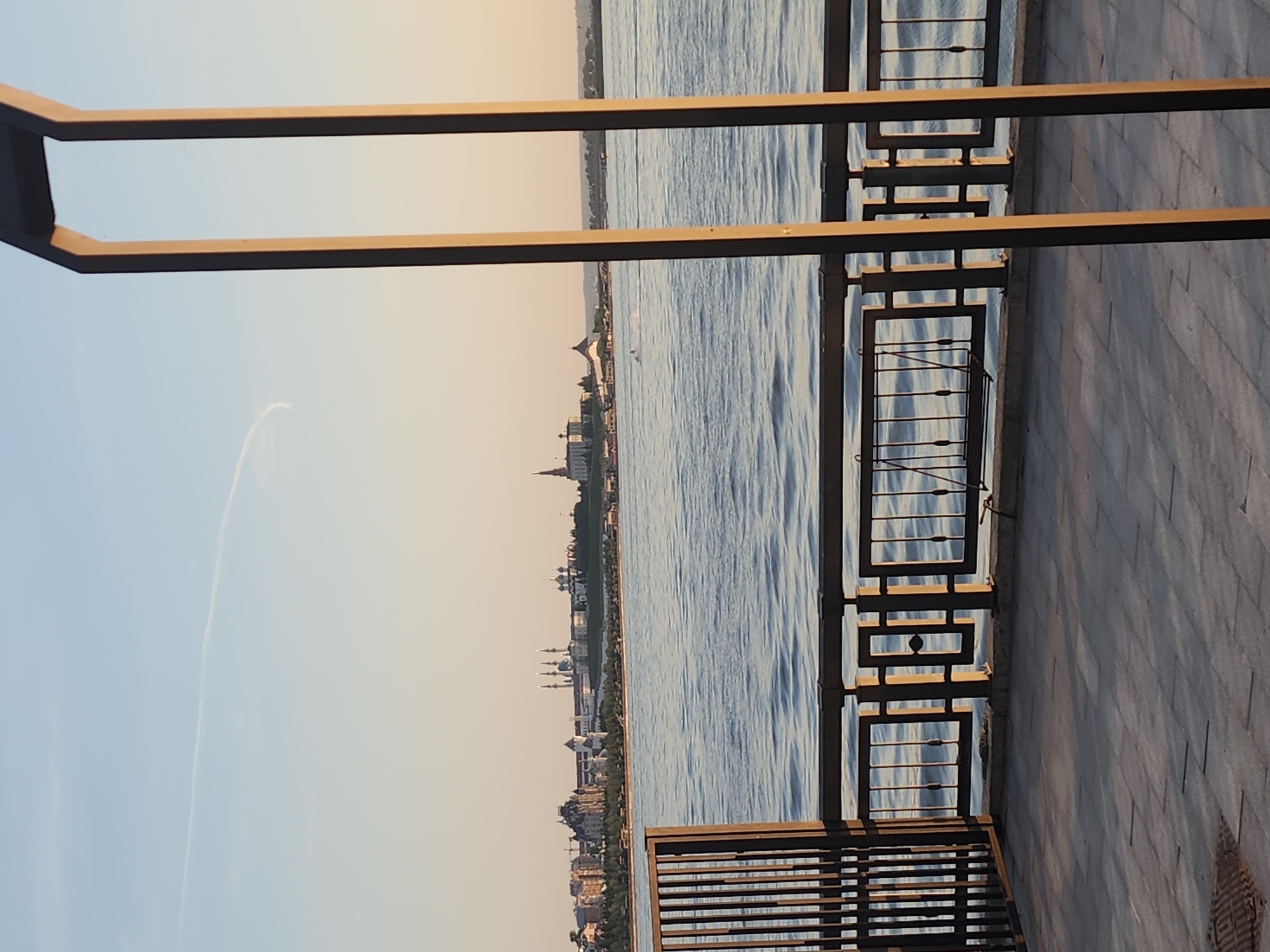 The image captures a view of a waterfront in an unknown city. The scene is framed by elements of a promenade, including a modern metal railing and parts of a seating structure in the foreground. Beyond these elements is a wide body of water reflecting the sky, which transitions from a pale blue to a subtle sunset hue near the horizon. On the opposite bank, a city skyline is visible, featuring a mix of architectural styles, including what appear to be domes and spires. The sky also contains a faint contrail, indicative of air traffic. The overall atmosphere is serene, captured during what seems to be either sunrise or sunset.