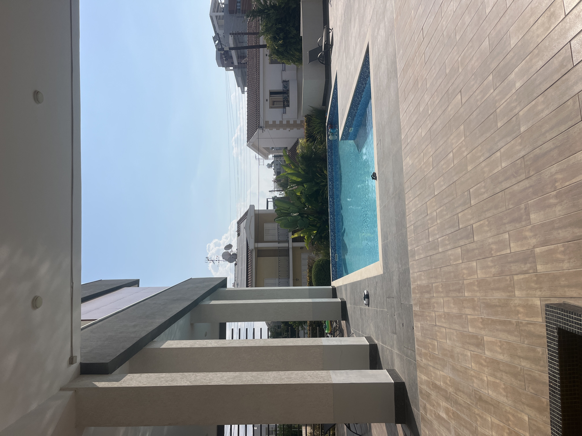The image depicts a modern outdoor swimming pool area adjacent to a building with large pillars, likely in Germasogeia Municipality, Cyprus. The pool is rectangular and filled with clear blue water, with a patterned tiled edge. The surrounding area is paved with large, light-brown wooden-look tiles. In the background, residential buildings with tiled roofs are visible, along with lush green foliage and trees. The sky is clear blue with a few scattered clouds. There are no people visible in the image, and the scene appears tranquil.