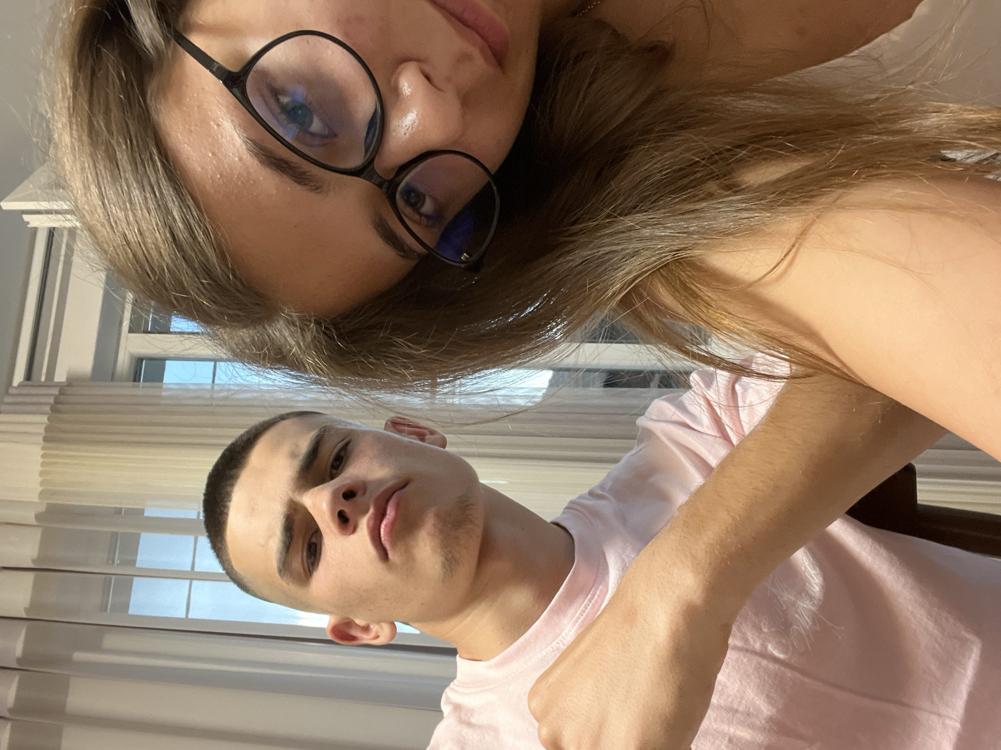 The image captures two individuals, one male and one female, in an indoor setting in Karavelovo, Bulgaria. The female is positioned to the right and closer to the camera, her face partially cropped on the right side. She has long, light brown hair that falls over her shoulders and obscures portions of the male. She wears black-framed eyeglasses with visible blue light reflections on the lenses, and her right eye is directed at the camera. Minor skin texture is visible on her forehead. The male is positioned to the left, slightly behind the female's shoulder and hair. He has a very short buzz cut and light stubble visible on his jawline. He wears a light pink crew-neck t-shirt with the words "Smiths Music" faintly visible in white text on the chest. His right arm is bent at the elbow, and his fist is lightly clenched and positioned in front of his body. Both individuals are looking directly at the camera. The background features interior windows draped with sheer white curtains, through which some daylight and hints of exterior blue are visible.